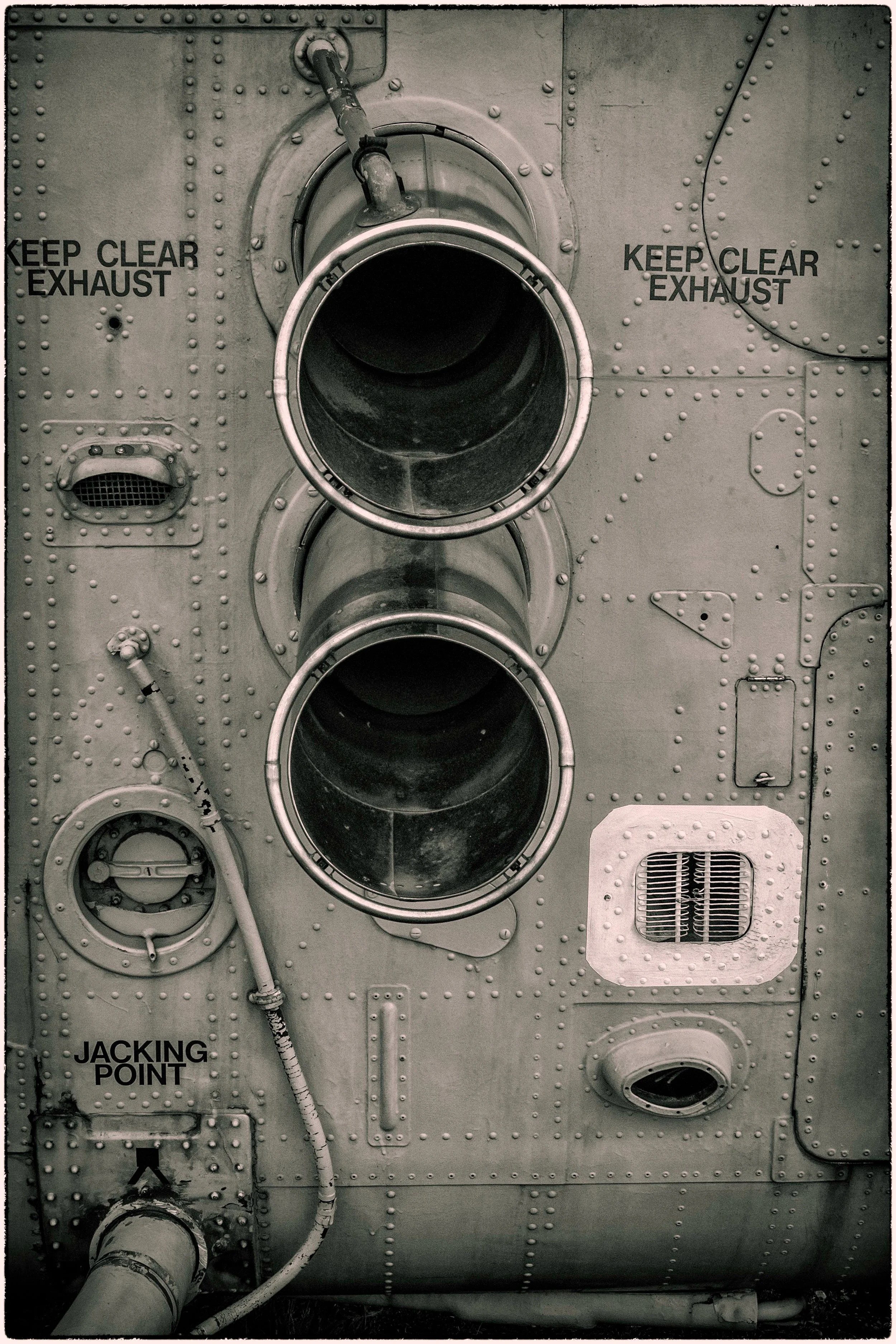 Close-up of an aircraft jet engine exhaust area with warning signs that say 'Keep Clear Exhaust' and 'Jacking Point' indicating safety zones and maintenance points.