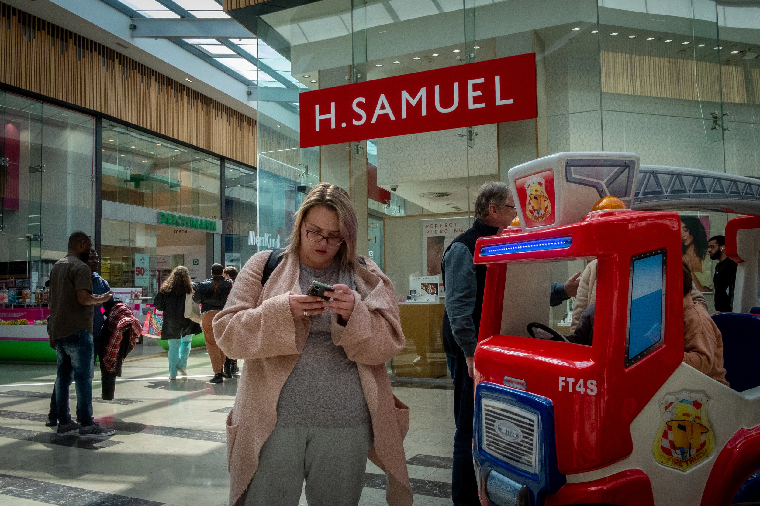 A woman in a pink coat looking at her phone in a shopping mall, with a red and blue toy truck in the foreground, and a red sign that reads 'H. SAMUEL' hanging from the ceiling.