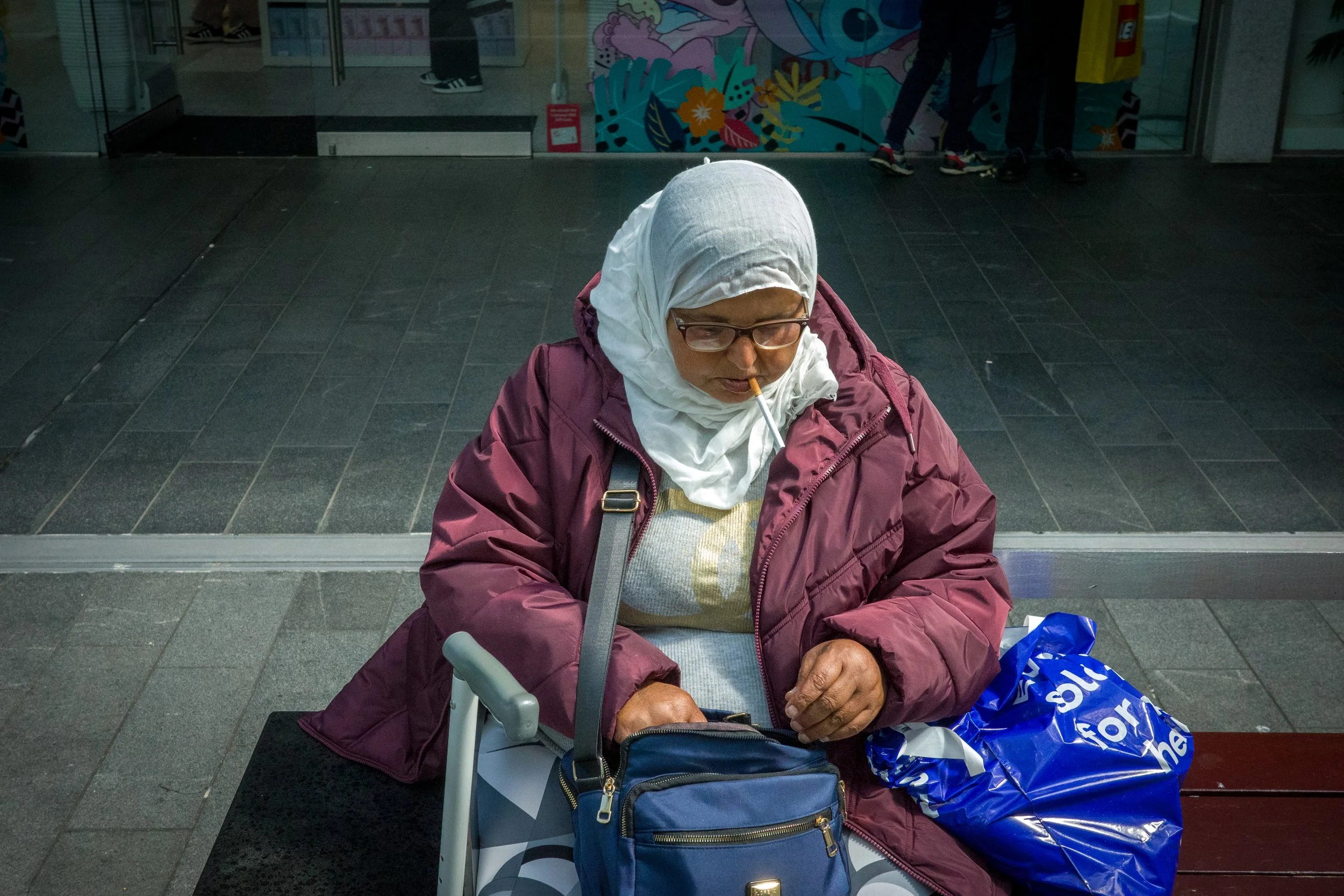 An elderly woman wearing a white hijab, glasses, and a maroon jacket sitting on a bench in an indoor public space, possibly an airport or train station. She has a cigarette in her mouth and is looking down at her hands. There are shopping bags, a sma