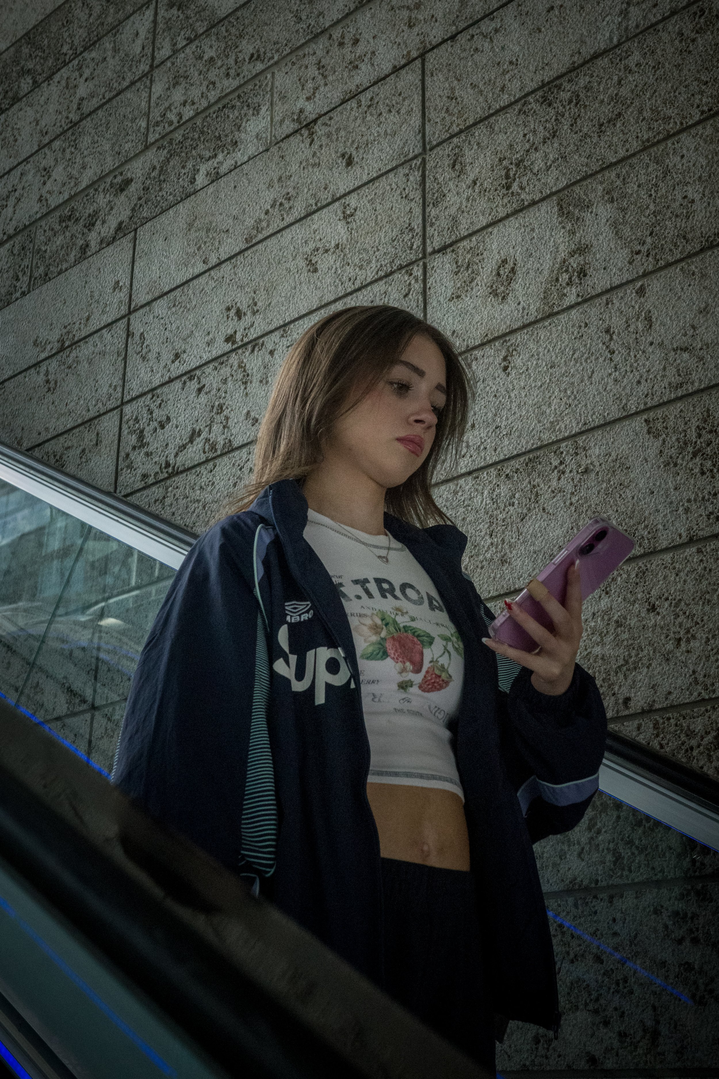 Young woman with long brown hair, wearing a black jacket over a white crop top with a fruit print, looking at her pink phone while standing on an escalator near a textured stone wall.