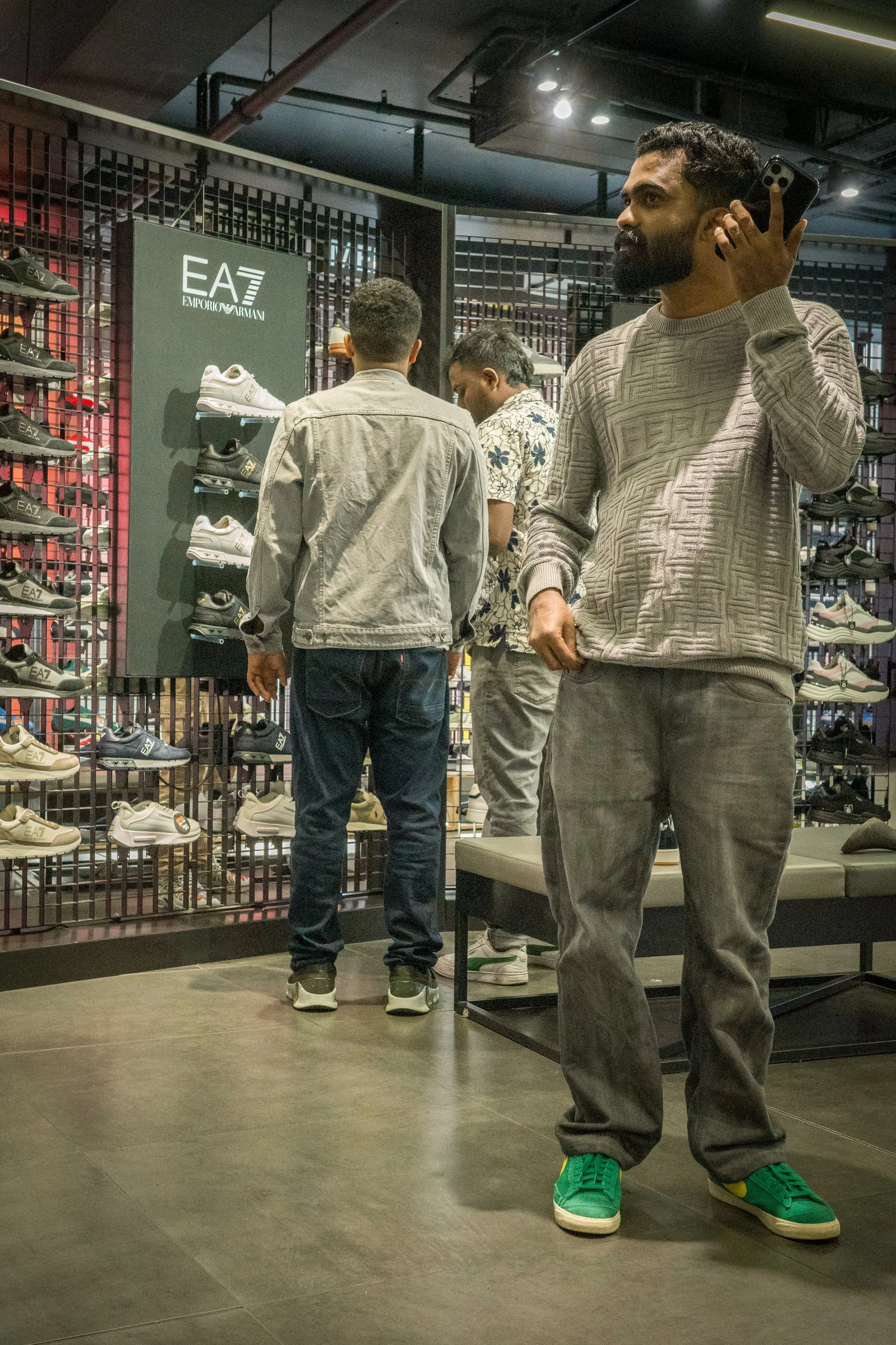 A man in a gray patterned sweater and jeans with green sneakers stands in a shoe store, talking on a cellphone, with other shoppers browsing Nike sneakers in the background.