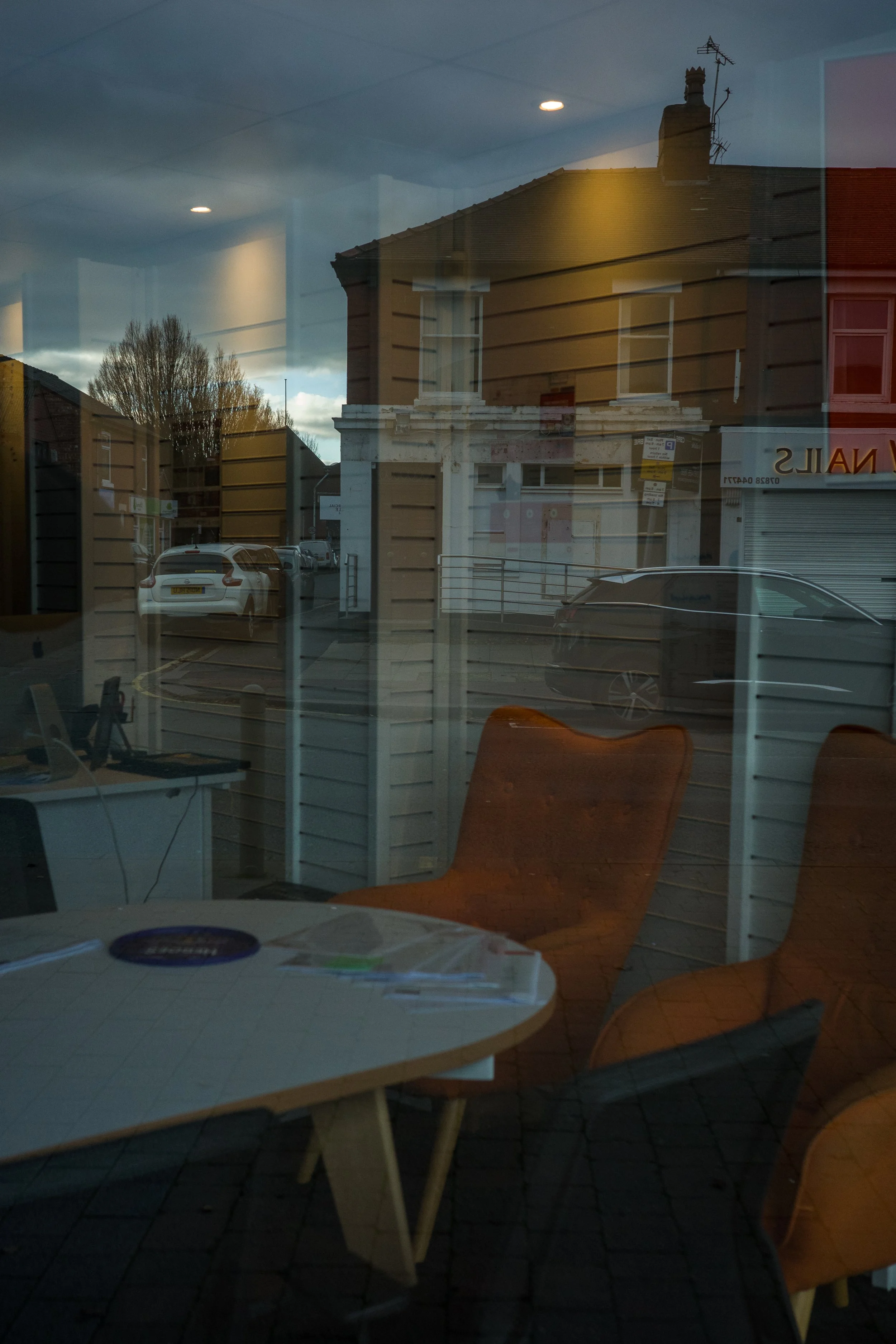 Reflections of parked cars and buildings outside on a glass window, with a table and orange chairs inside the room.
