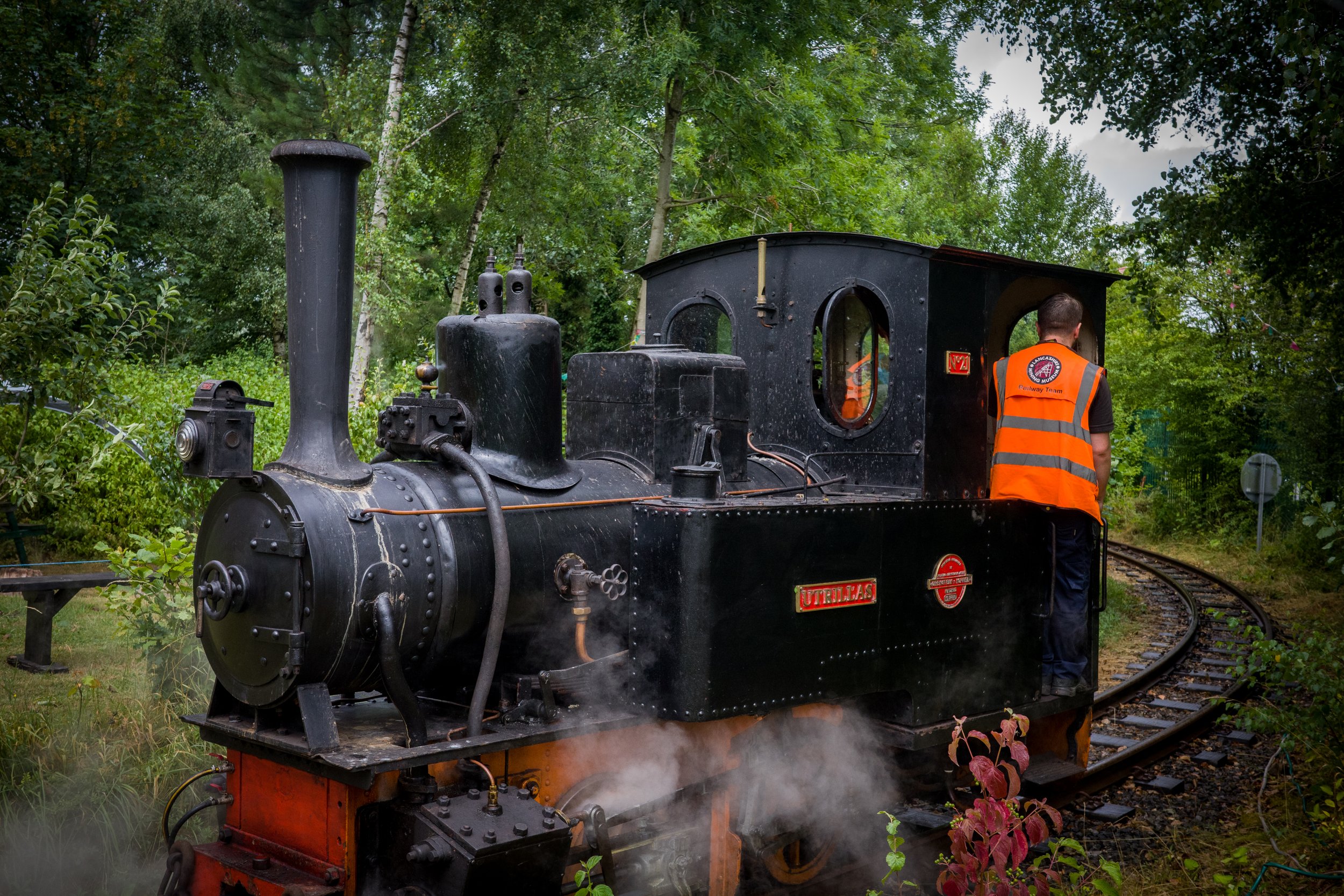 A black vintage steam locomotive on railway tracks surrounded by green trees and bushes, with a person wearing an orange safety vest standing near the engine.