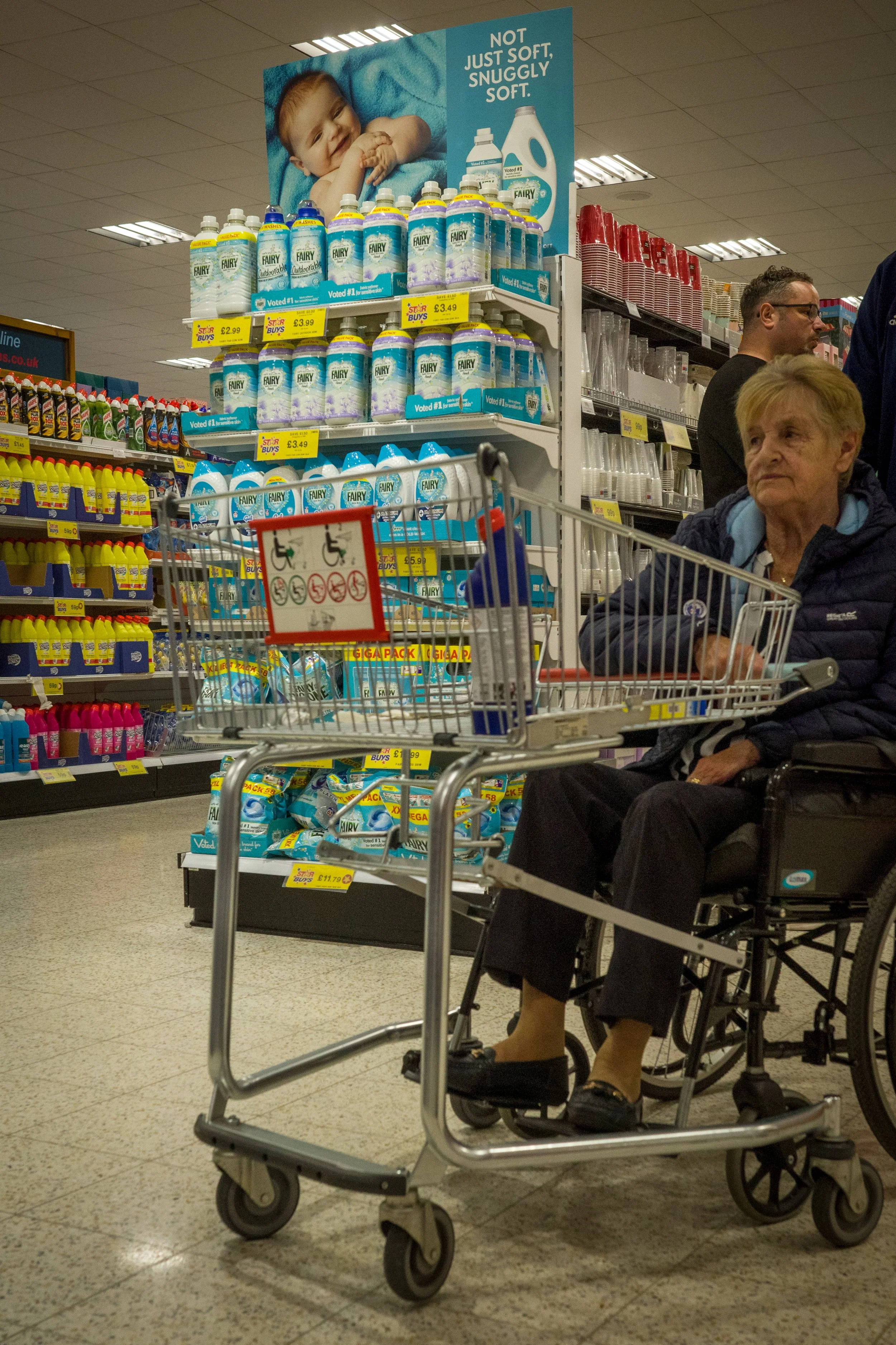 An elderly woman sits in a wheelchair at a store aisle with a shopping cart. The shelf behind her displays bottles of Fairy dish soap and a large sign with a baby and laundry detergent bottles. Other shoppers are visible in the background.