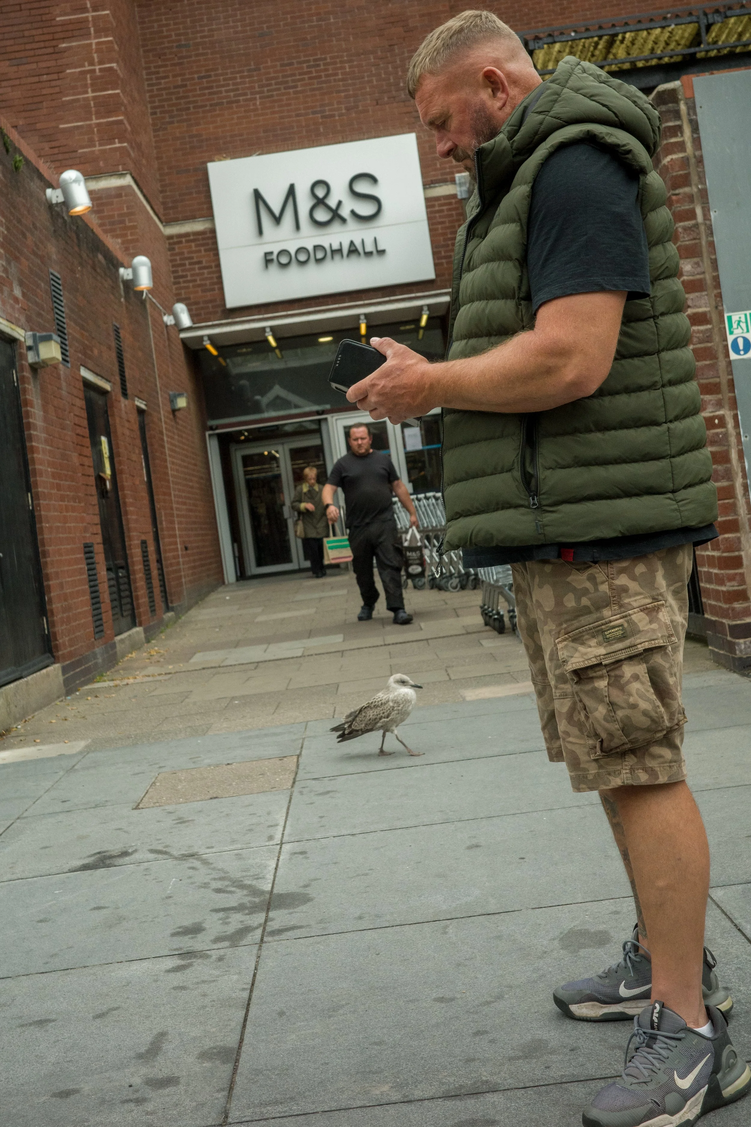 Man in camouflage shorts and grey sneakers stands on city sidewalk, looking at cellphone, with seagull nearby. In background, people walk past entrance of M&S Foodhall store.
