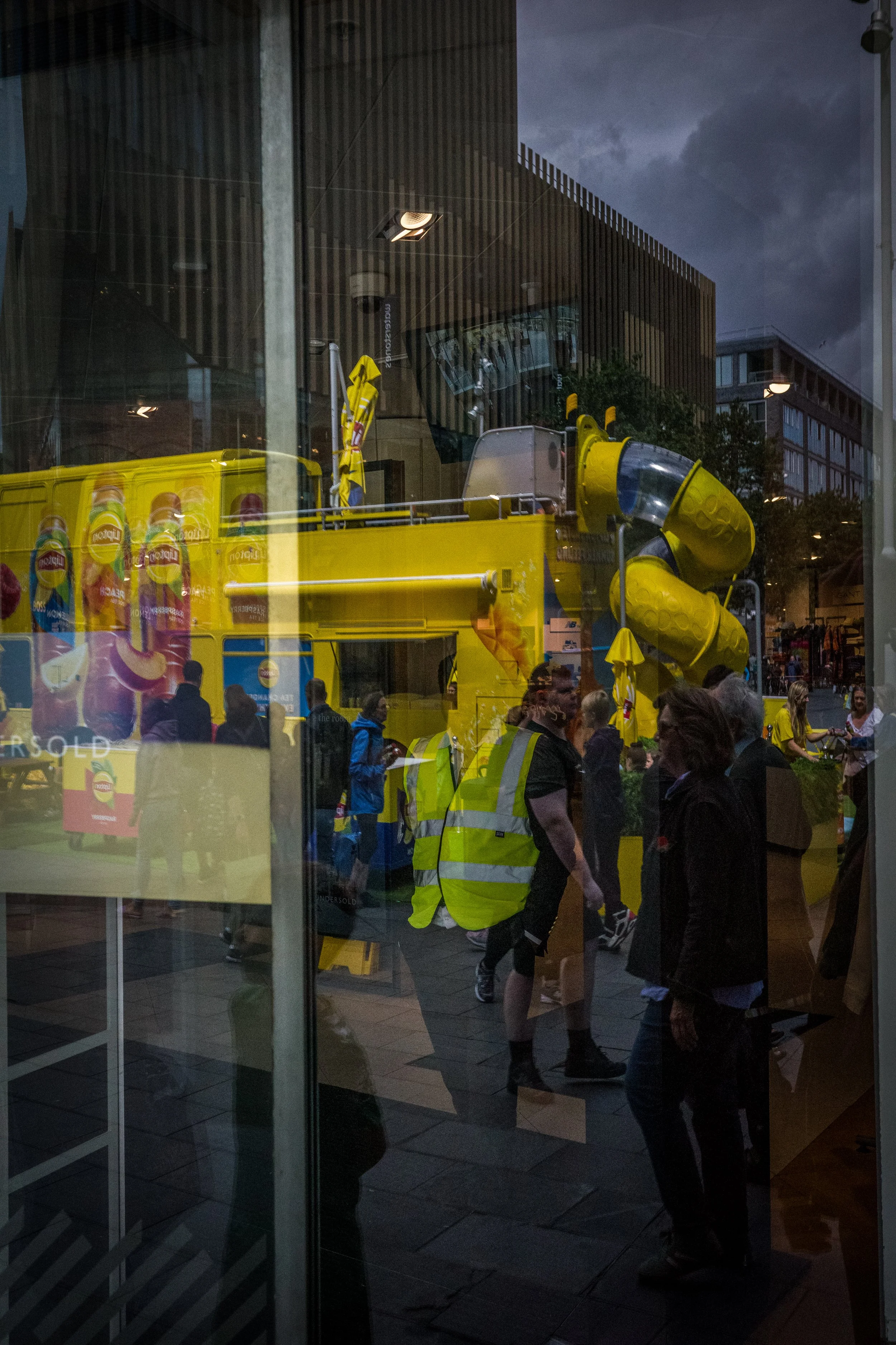 Reflection of a street scene with people and a yellow slide in a shop window during evening.