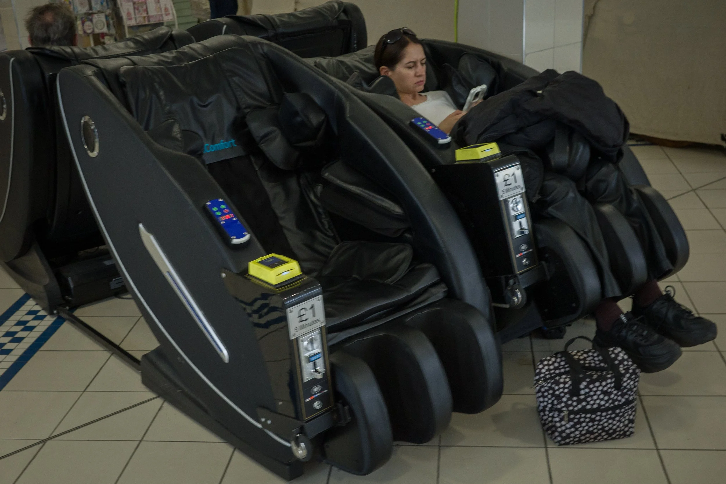 A woman sitting in a massage chair at an airport, using her phone with headphones nearby. There is a small yellow device and a pack of gum on the chair's armrest. A child's polka-dotted bag is on the floor next to her.