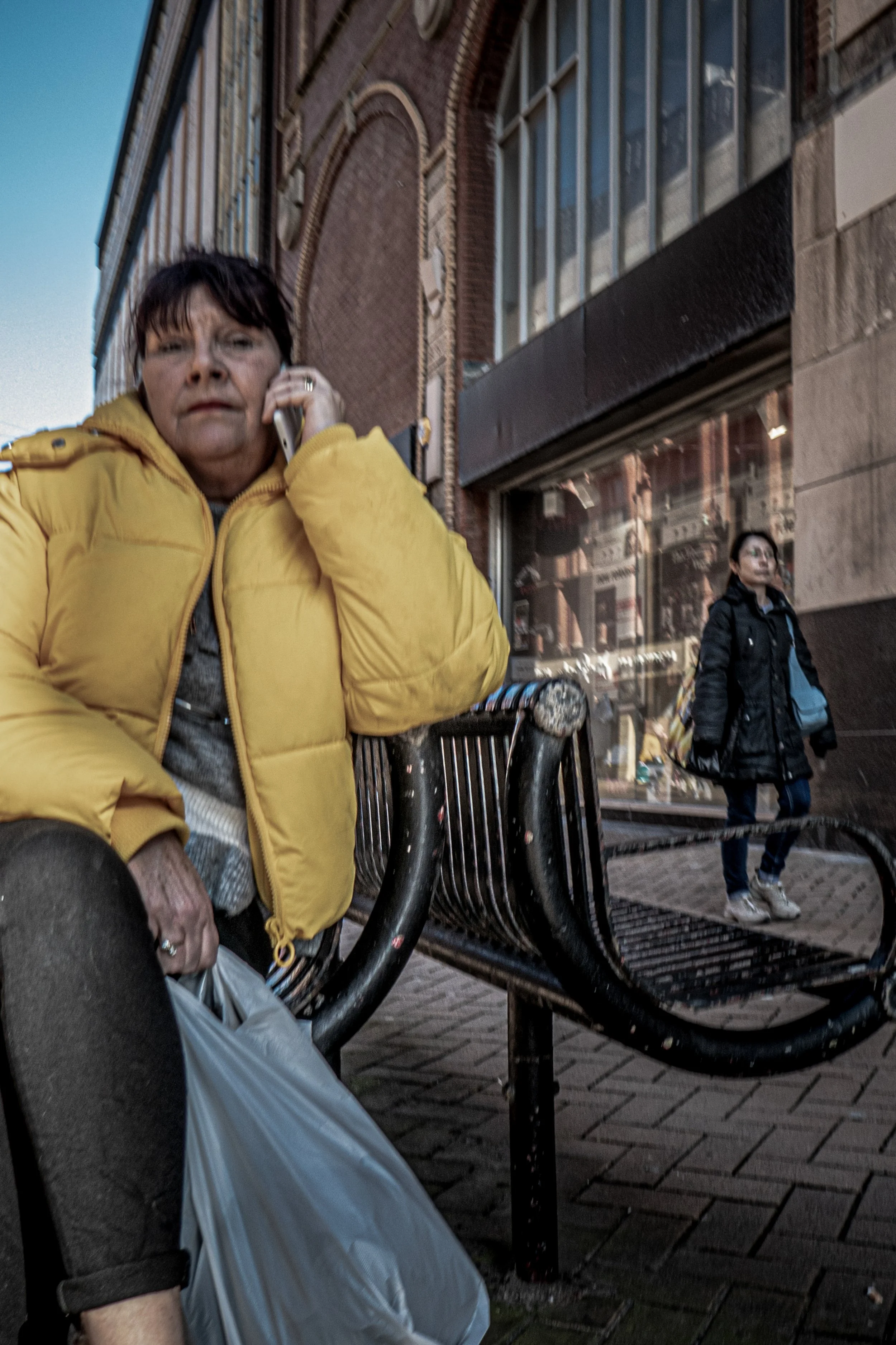 A woman in a yellow jacket talking on her cellphone, sitting on a bench on a city street, with another person walking by in the background, charred bike racks, and tall brick and glass buildings.