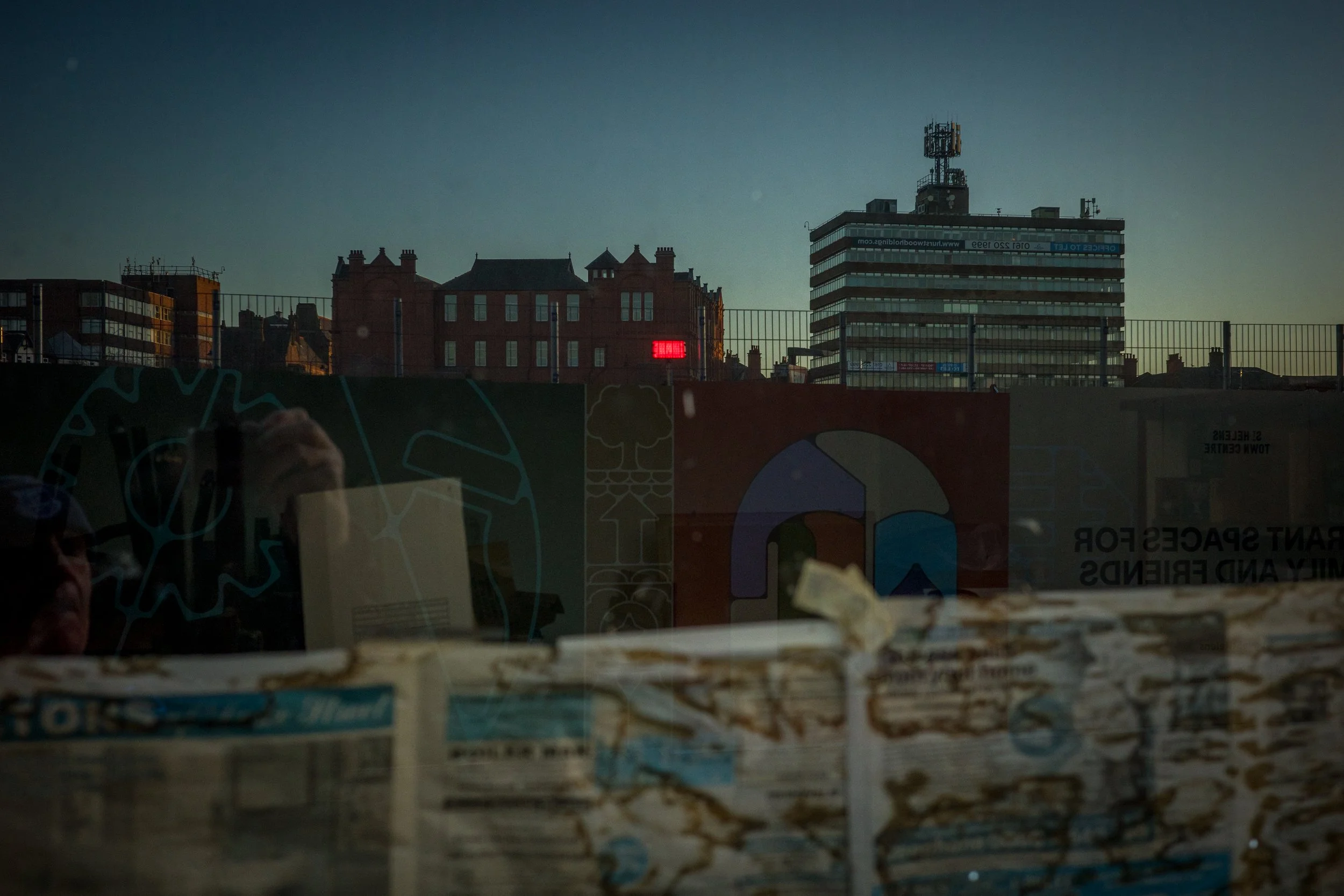 City skyline during sunset with buildings, some with illuminated signs, viewed through a glass window that reflects the interior including a person taking a photo, newspaper, and some colorful artwork.