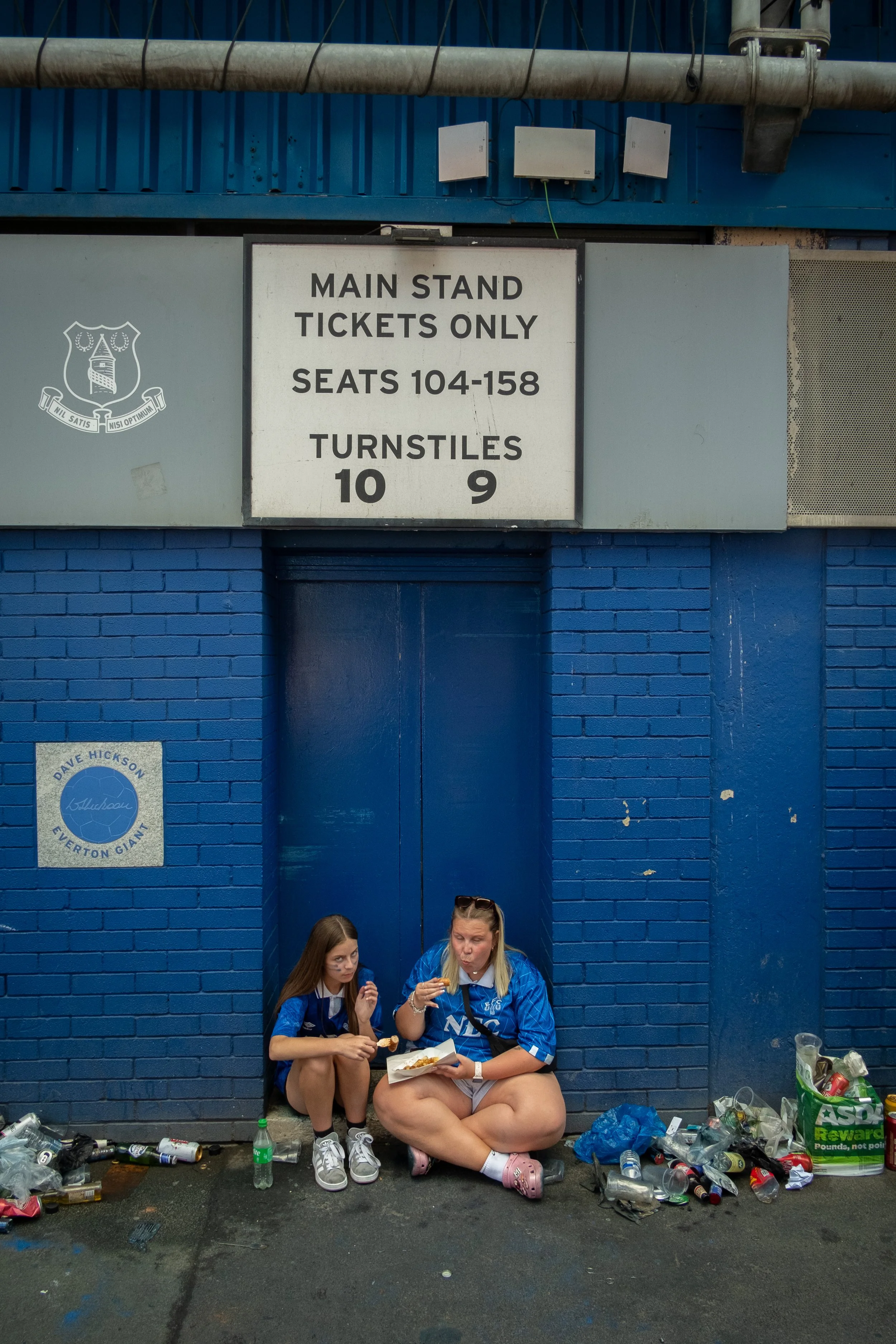 Two young women sitting on the ground outside a sports stadium, eating food and surrounded by empty bottles and trash, underneath a ticketing sign indicating ticket sections 104-158 and turnstiles 9 and 10.