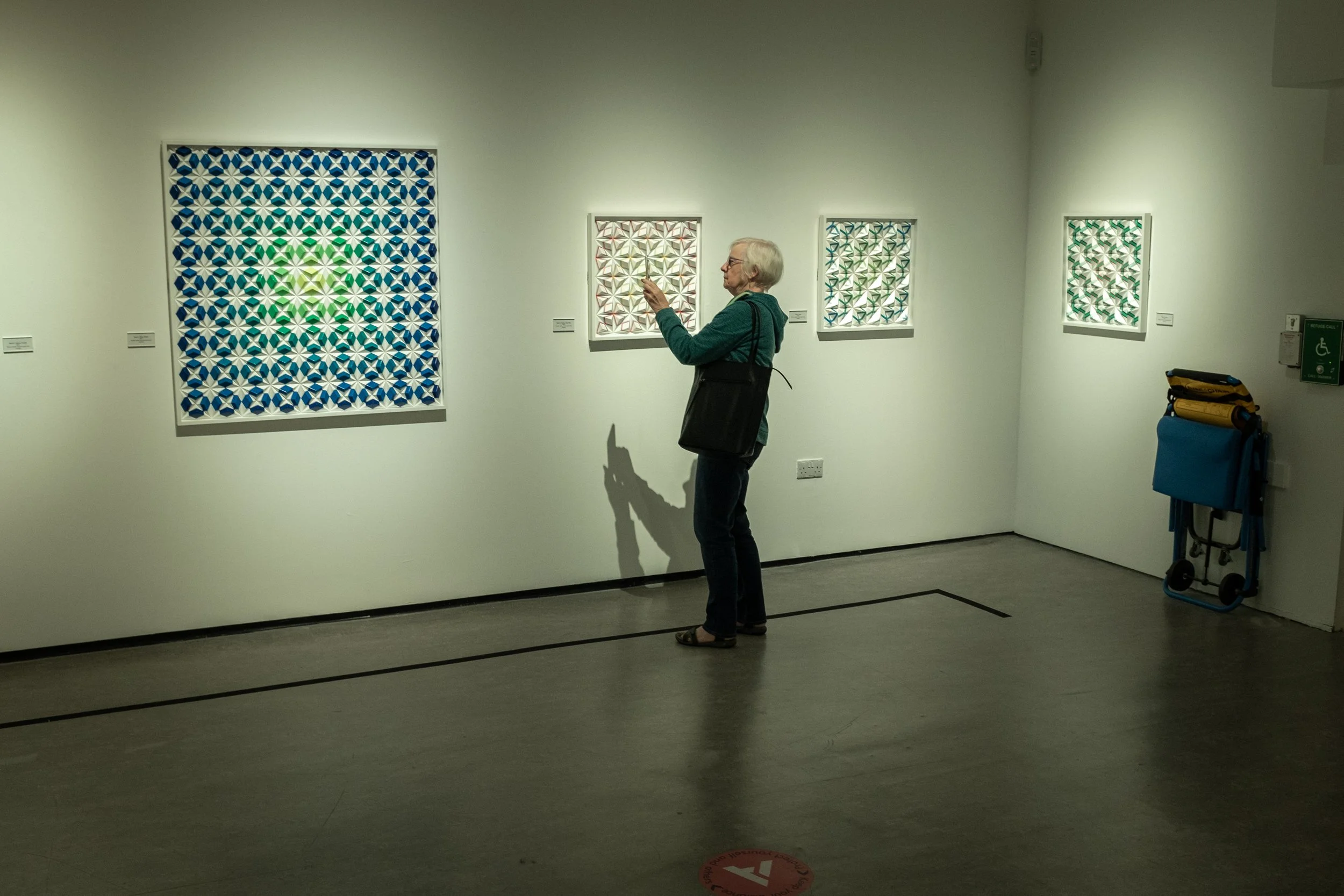 An elderly woman with white hair and glasses, wearing a green jacket, is standing in an art gallery looking at a colorful, geometric-patterned artwork. She is holding her phone and has a black bag on her shoulder. There are four framed pieces of art 