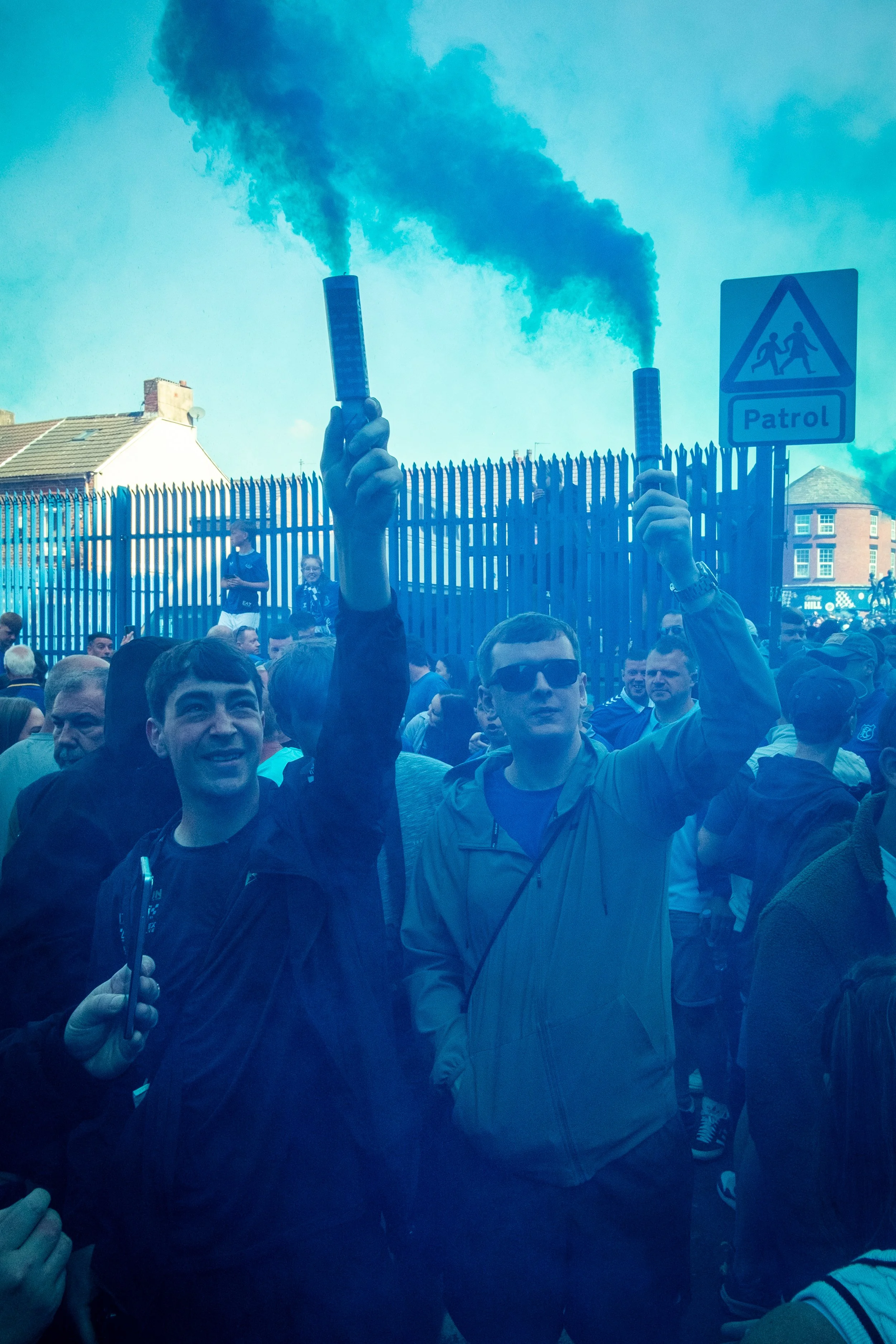 Two men holding smoke flares at a crowded outdoor event or protest, with many people around and a sign that says 'Patrol', in front of a wooden fence and blue sky.