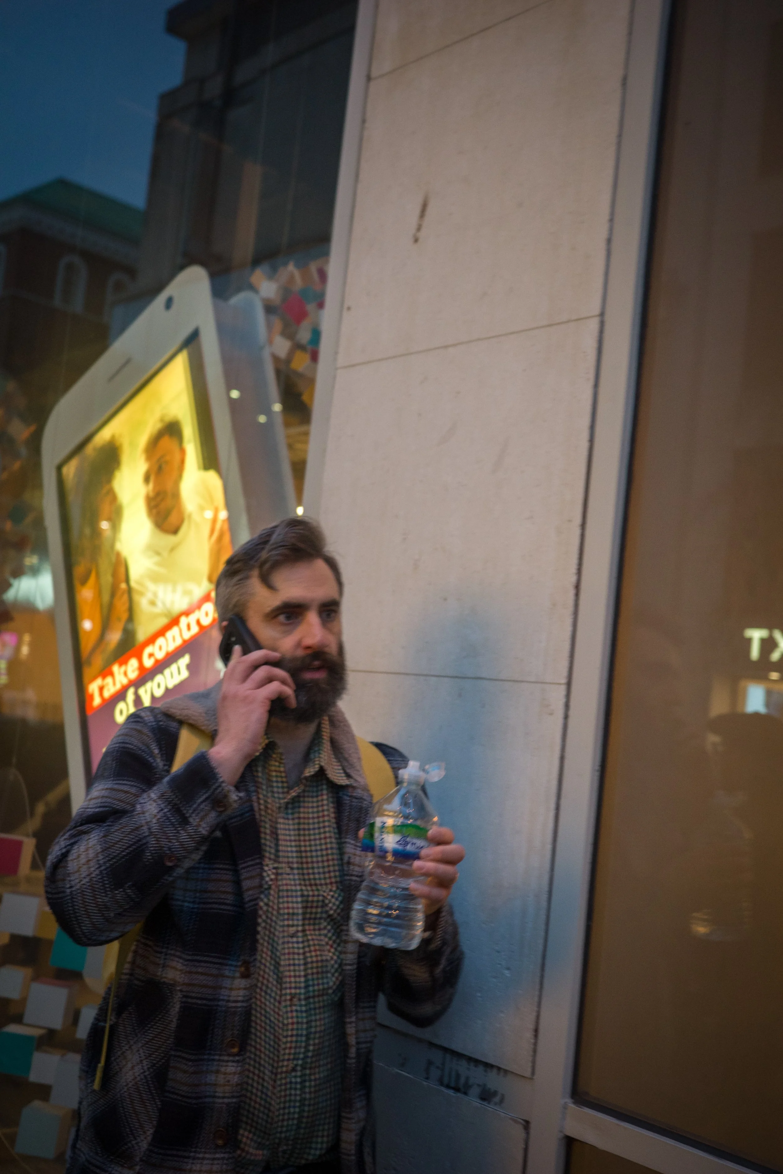 A man with a beard, wearing a plaid shirt and a jacket, is holding a water bottle in one hand and a phone to his ear with the other hand, standing outside near a wall with a large digital display and an advertisement board.