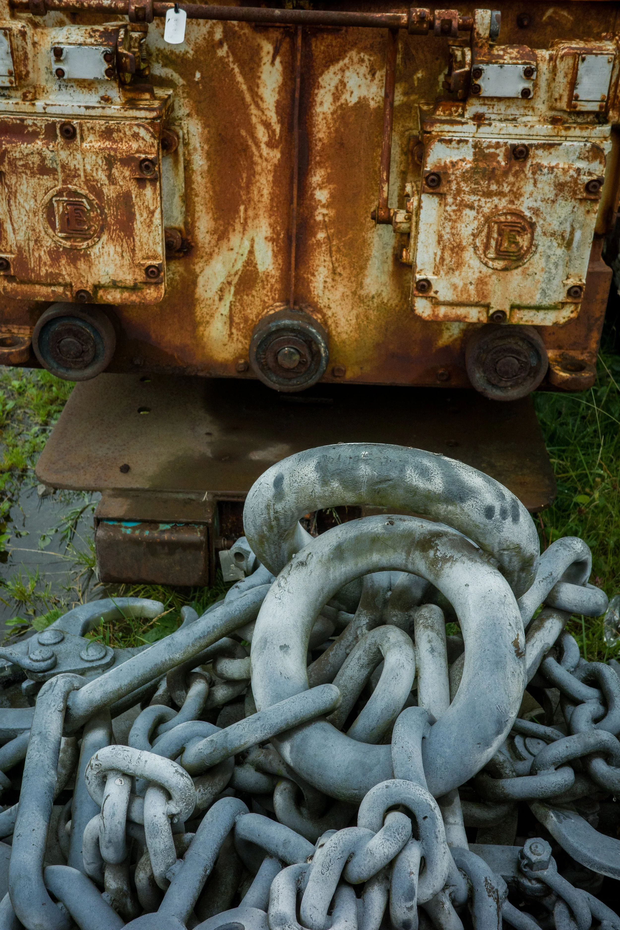 Rusty industrial machinery with two large control boxes mounted on a rusty metal surface, surrounded by coiled metal chains on the ground, outdoors.