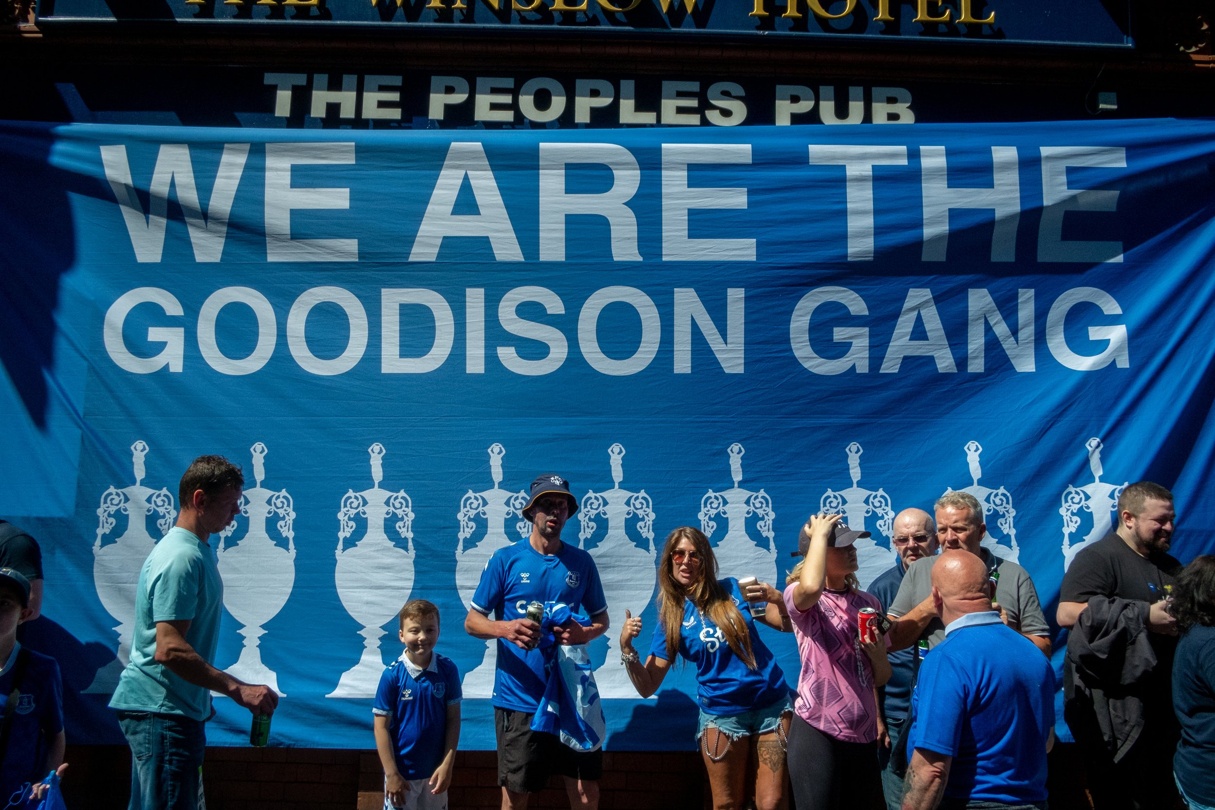 Group of people standing in front of a large blue banner that reads 'We Are The Goodison Gang' outside a pub.