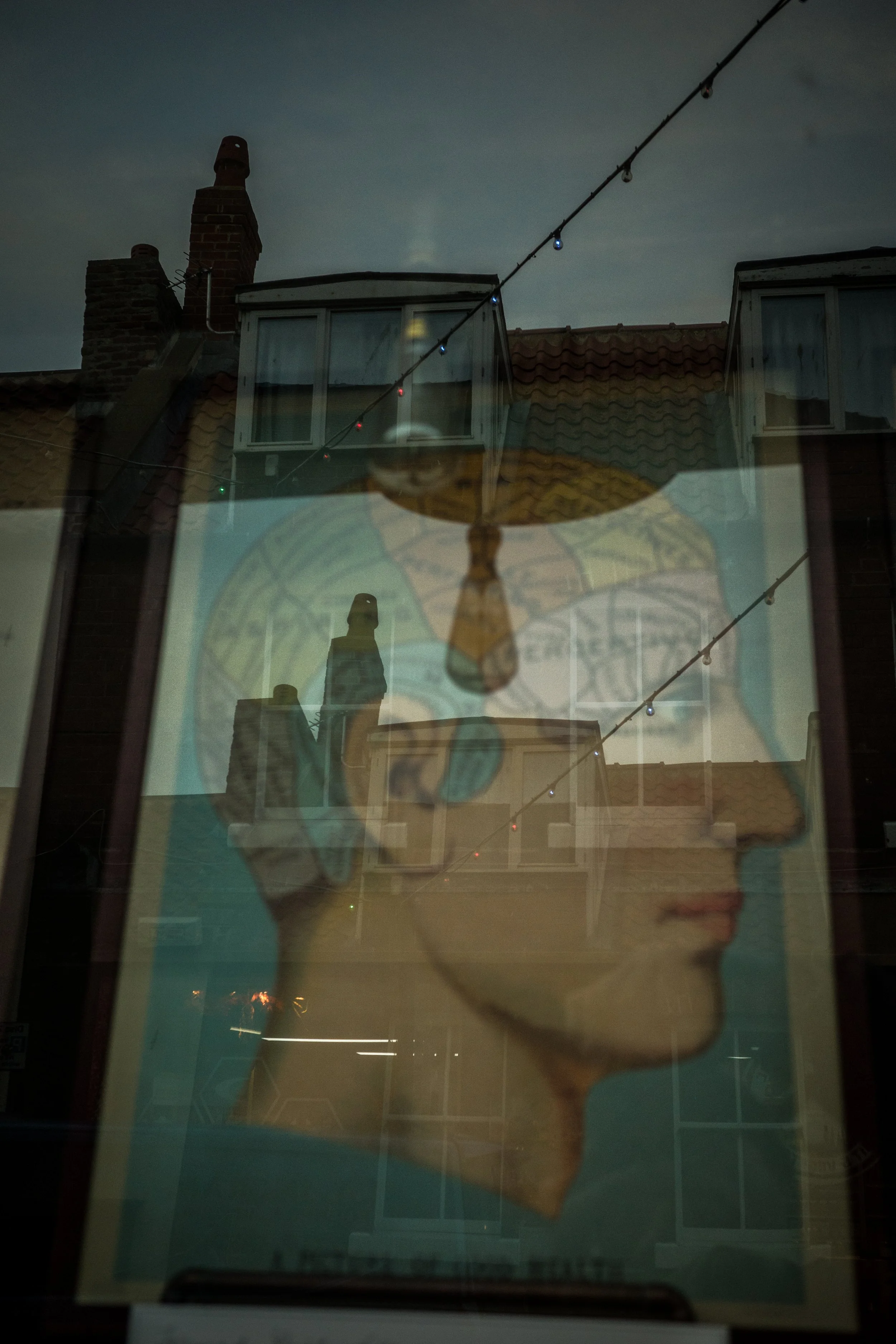 Reflection of a building with chimneys, windows, roof tiles, a string of lights, and a poster of a woman's face wearing glasses and a hat in a glass window.