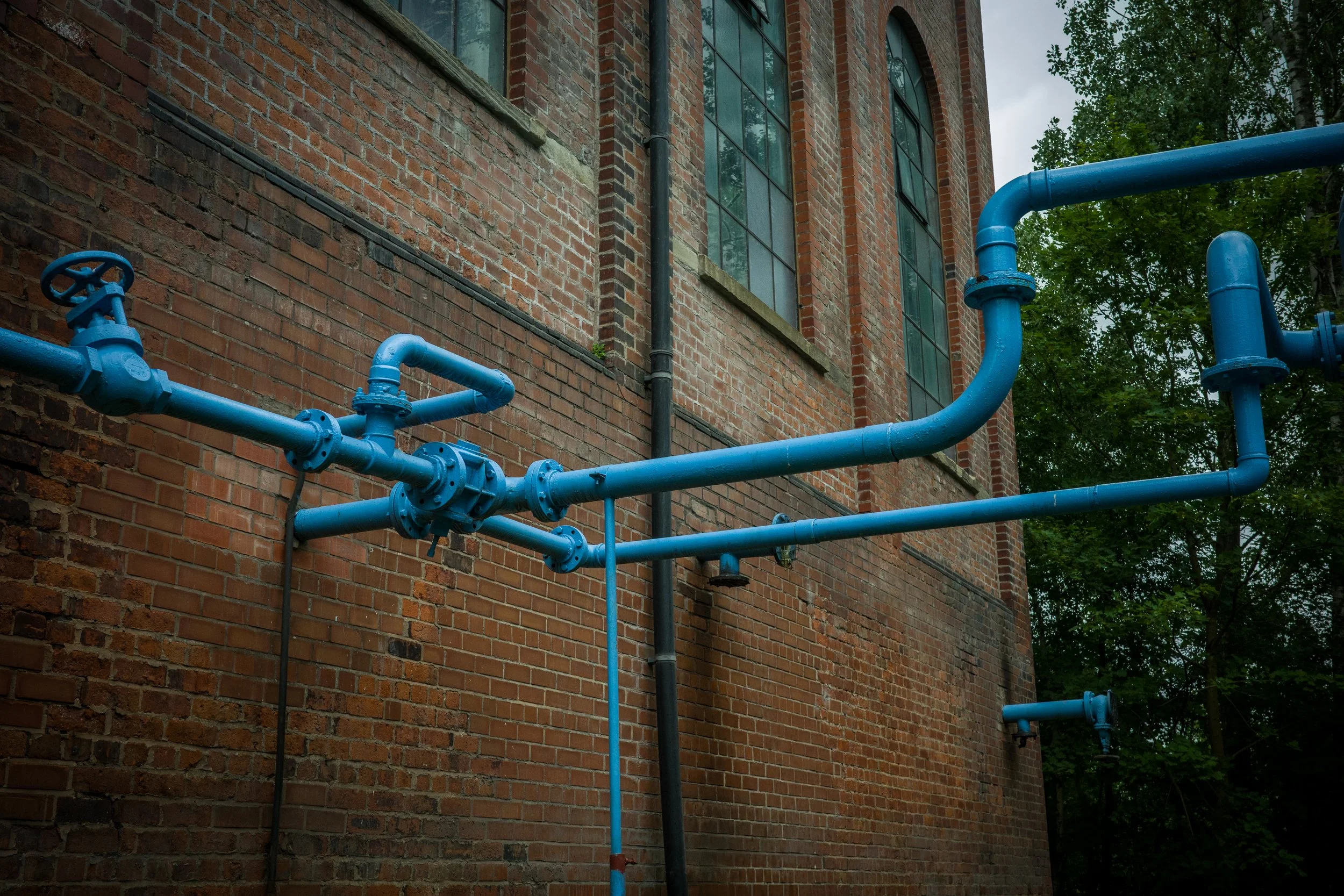 Blue industrial pipes running along the exterior brick wall of a building with large windows, green trees, and an overcast sky in the background.