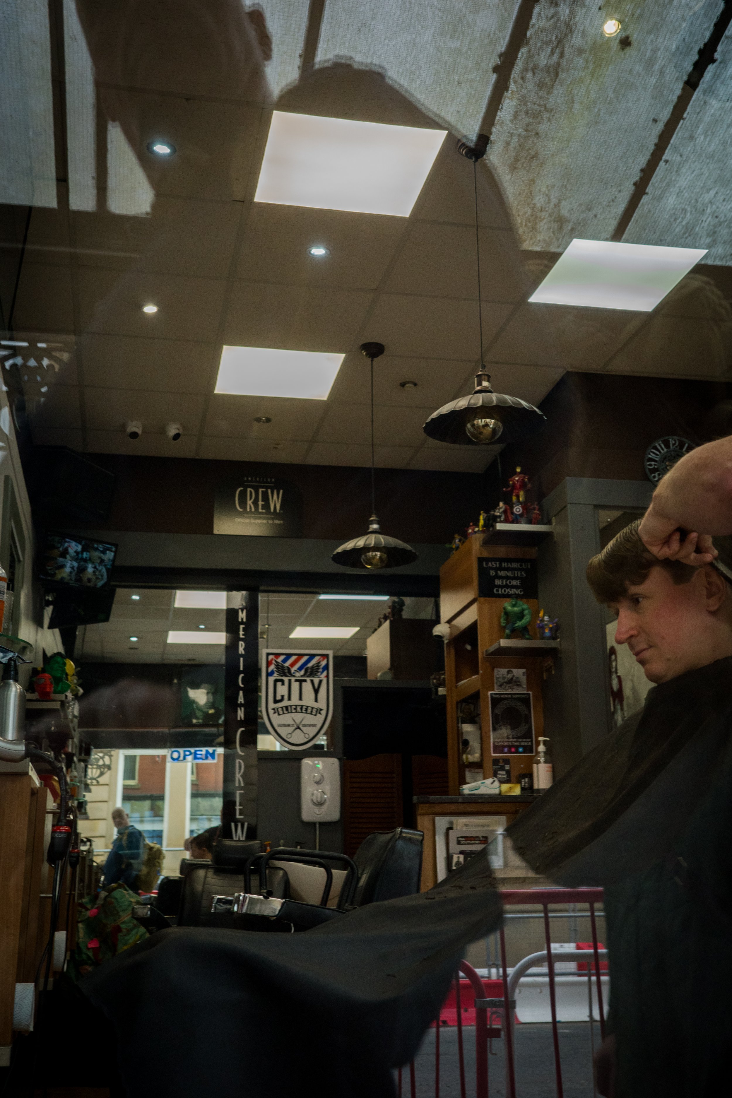 A man sits in a barbershop chair inside a barber shop, with a hand on his head, getting a haircut. The shop has posters, bottles, and other items on shelves, with a window showing pedestrians outside.