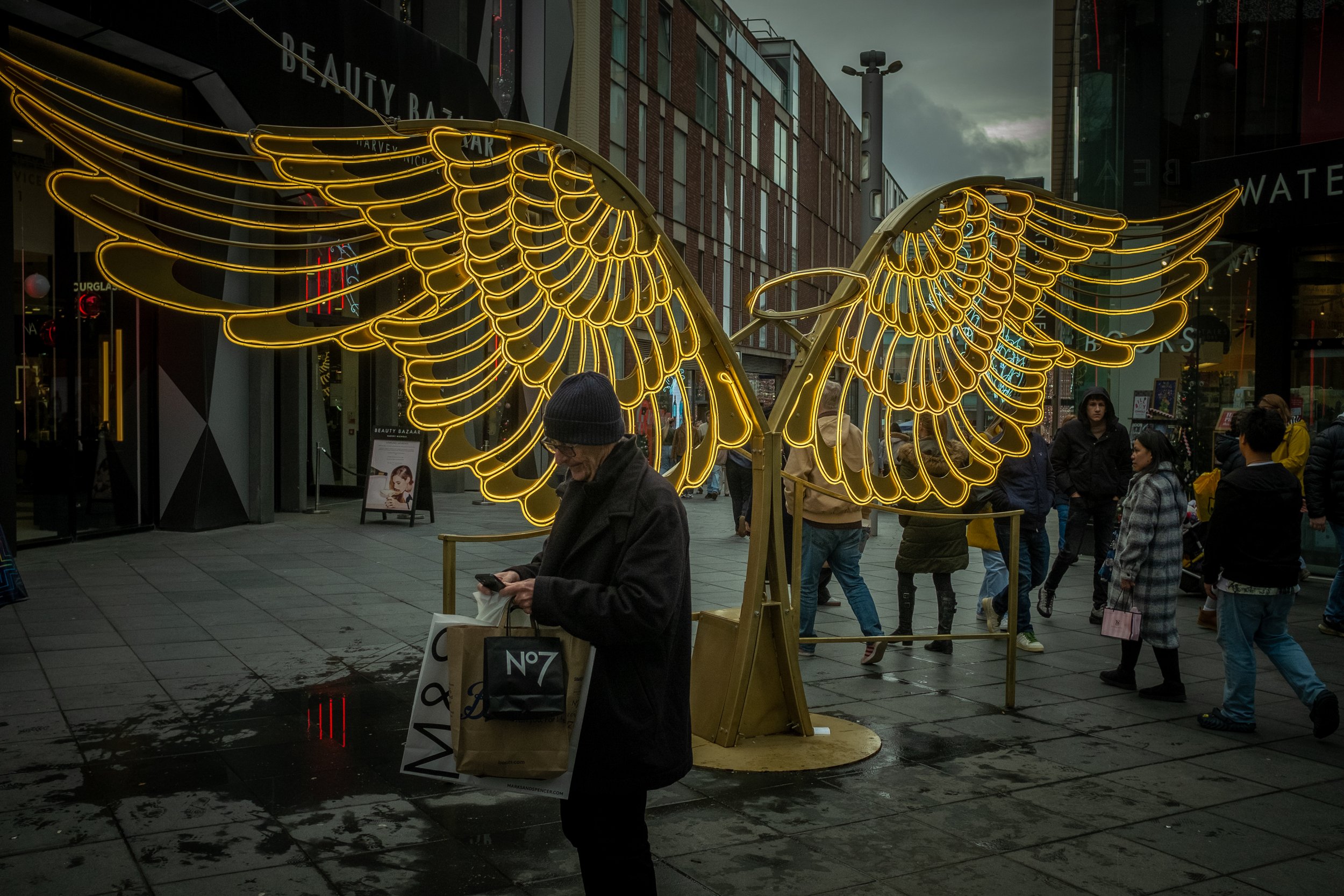 Illuminated golden angel wings display with a crowd of people walking by on a city street during evening or cloudy weather.