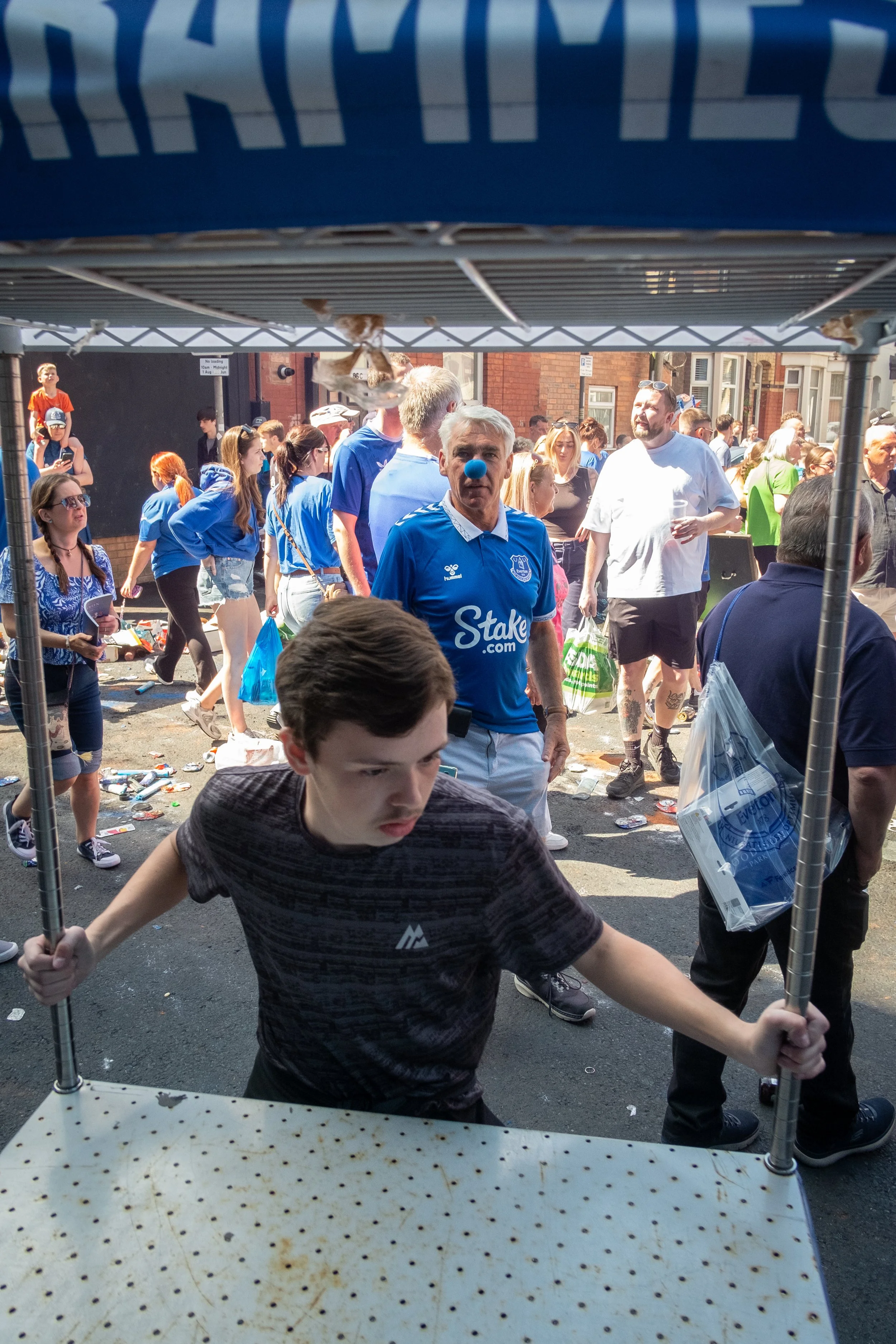 A young man with dark hair carrying a folding table with a speckled top while wearing a black athletic shirt, standing inside a metal-framed booth at a crowded outdoor event on a sunny day. Behind him, many people are walking and socializing, some we