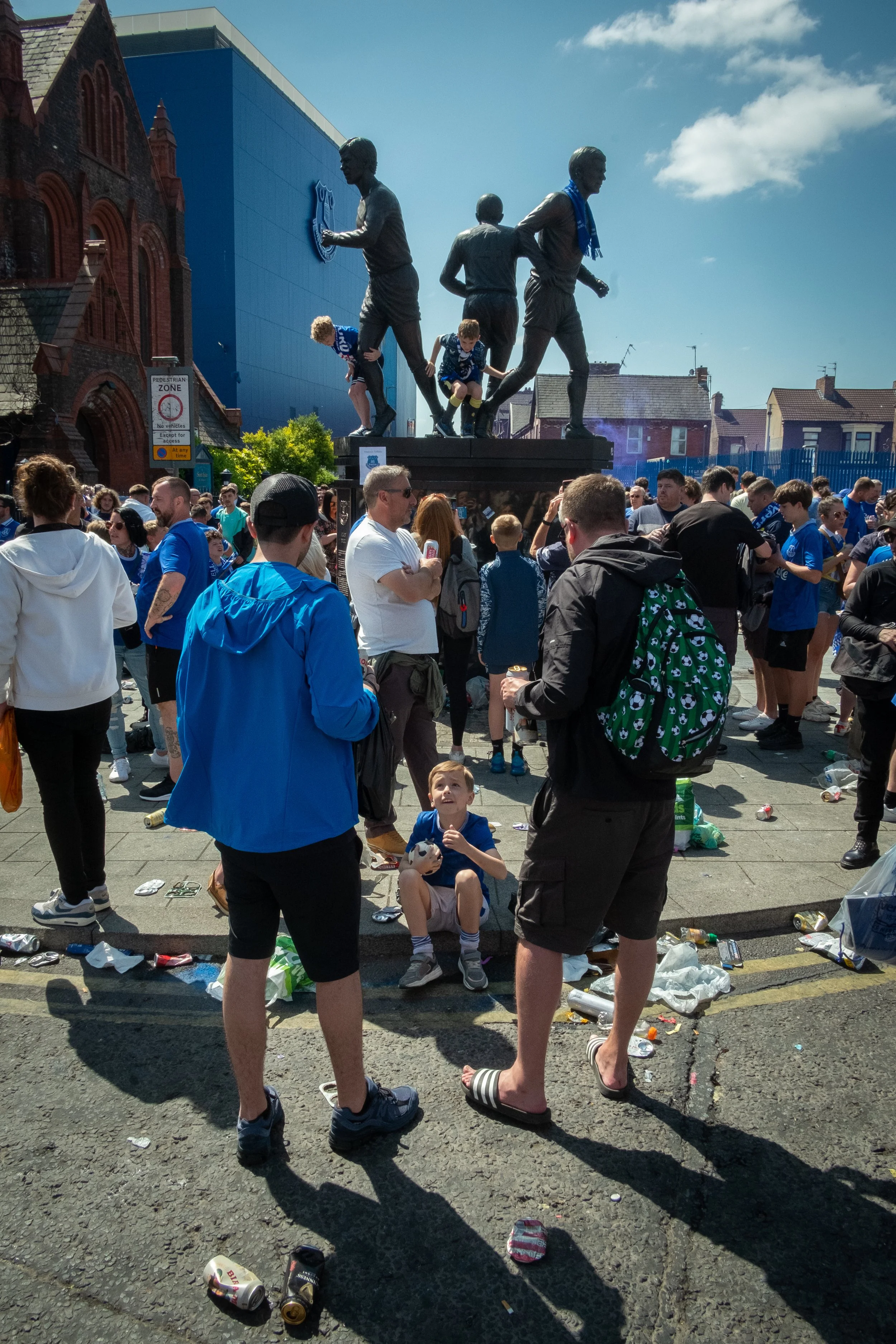 A crowd of football fans gathers around a statue of football players outside a stadium, with some climbing on the statue. Many are wearing blue jerseys, and the area is littered with trash on the ground. It appears to be after a game or event.