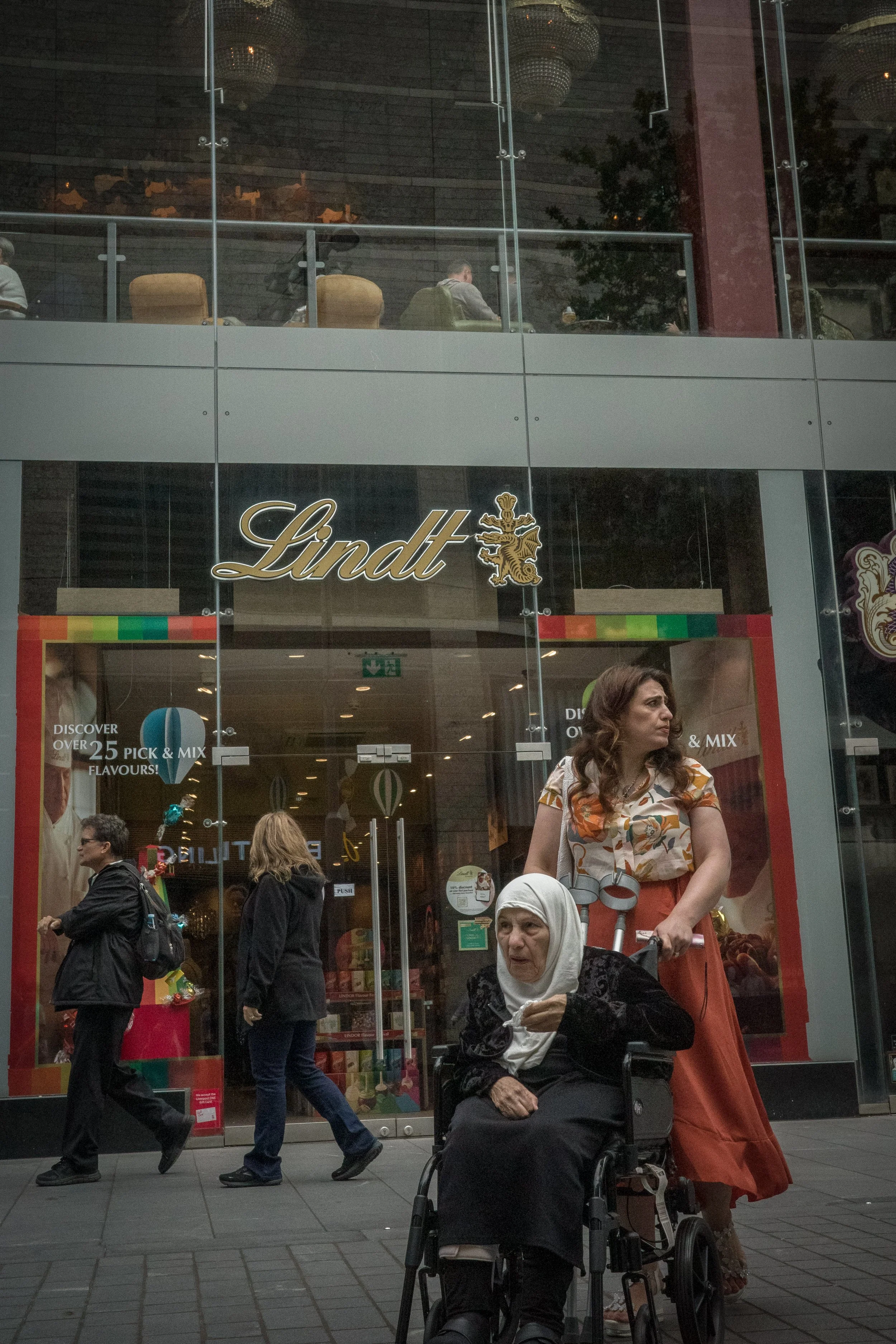 Street scene outside a Lindt chocolate store with several pedestrians, including a woman pushing a woman in a wheelchair, and an upper glass retail space with customers.