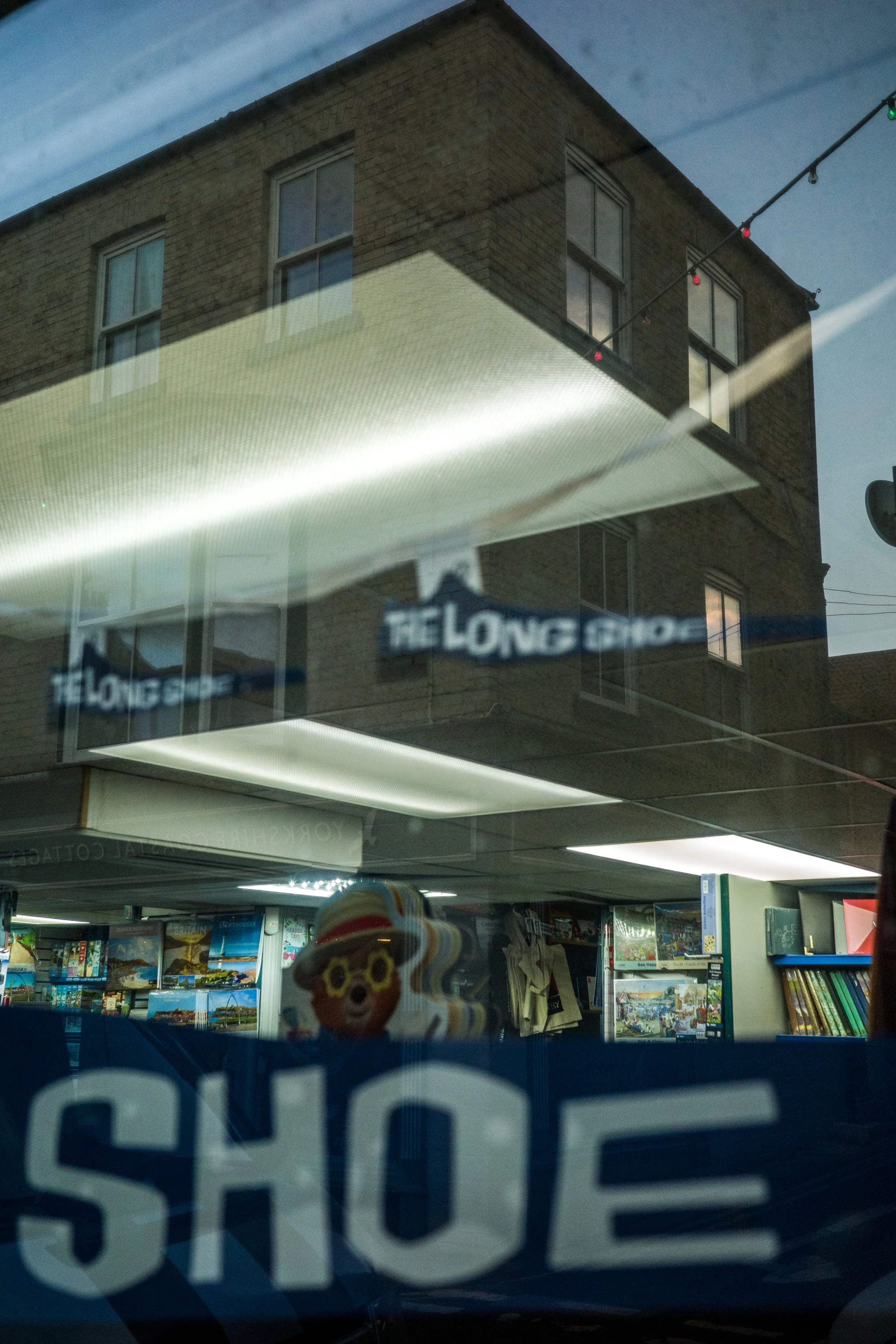 View through a store window showing a reflection of a brick building with windows and string lights, and the store interior with books and a decorative character sticker.