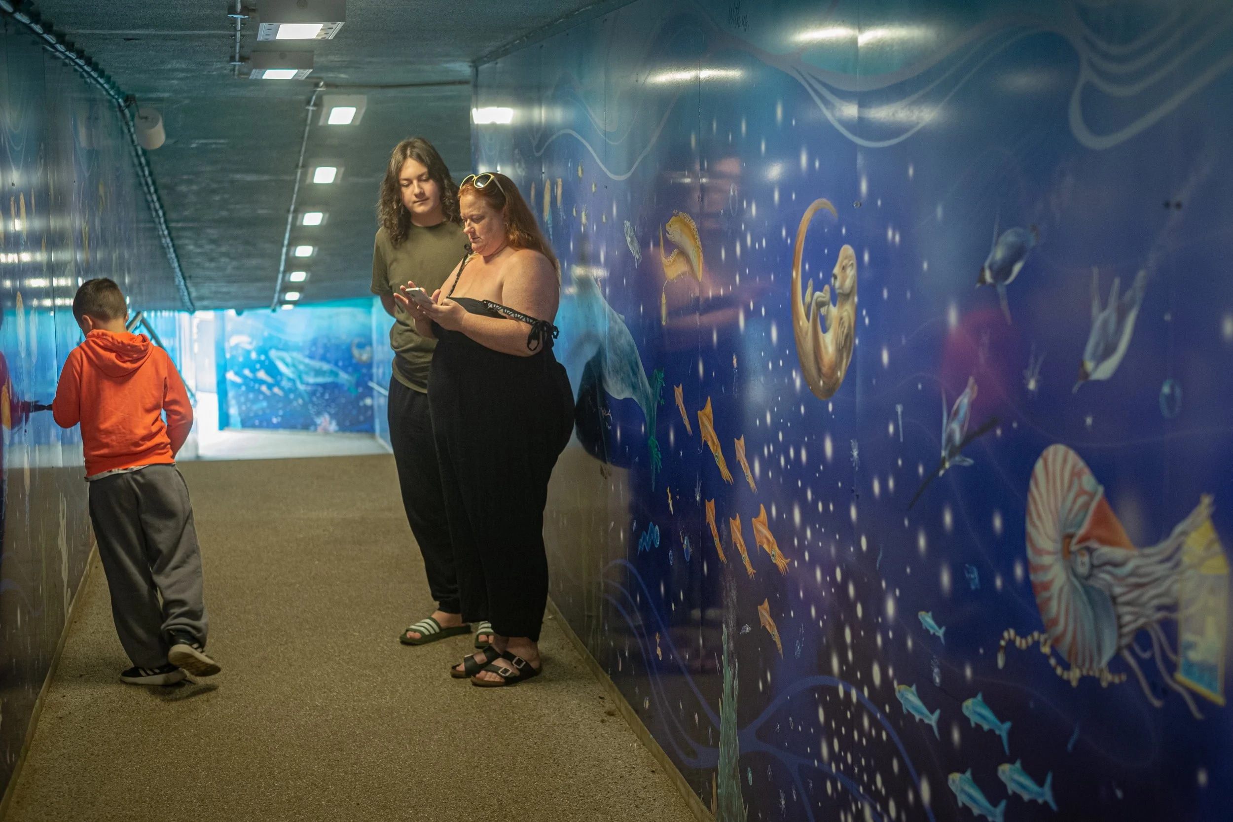 Three people standing and walking along a hallway with an aquatic mural featuring fish and sea creatures on the wall.