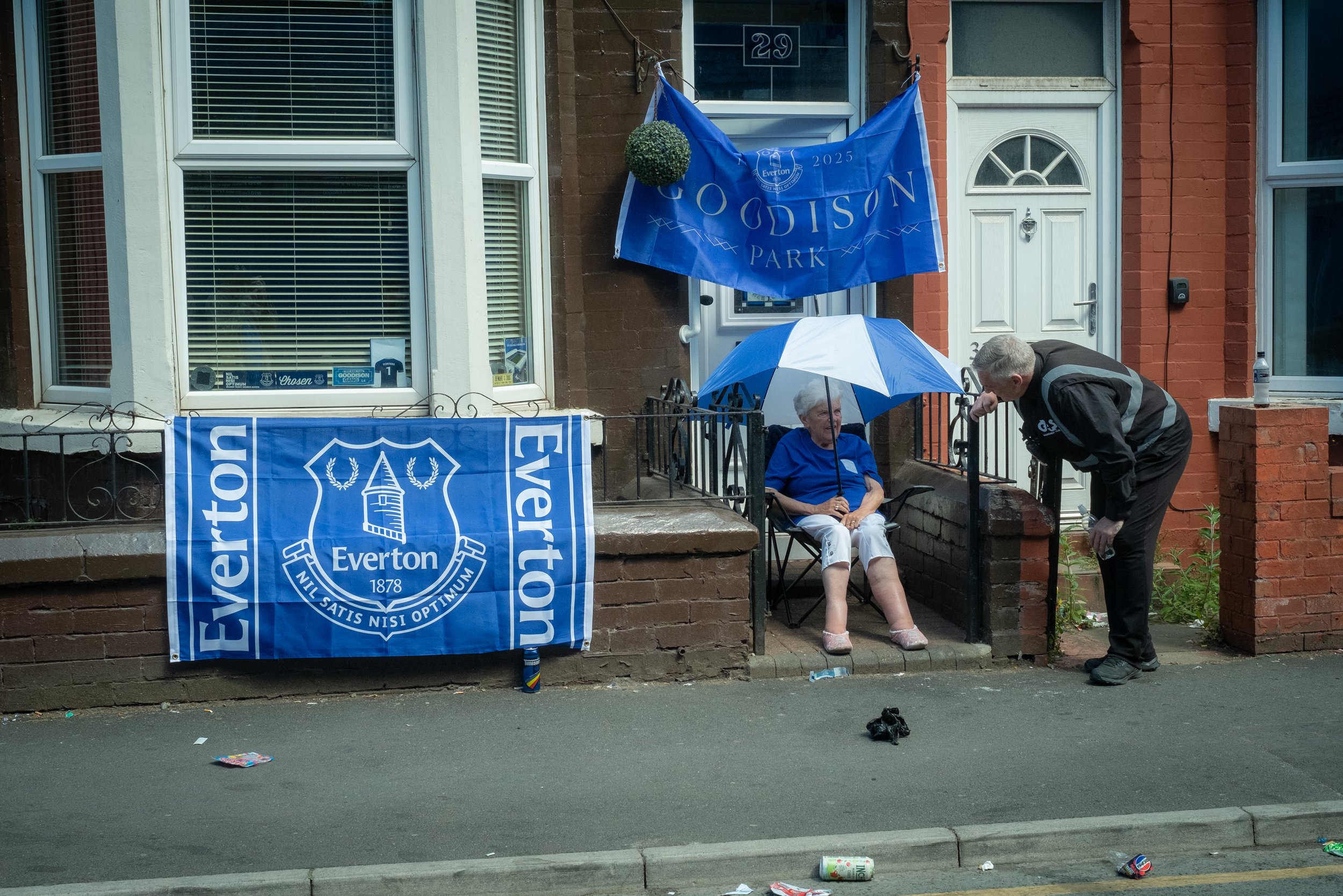 An elderly woman sitting on her porch with a blue and white umbrella, talking to a man. There are Everton flags hanging on her house, indicating support for Everton Football Club.