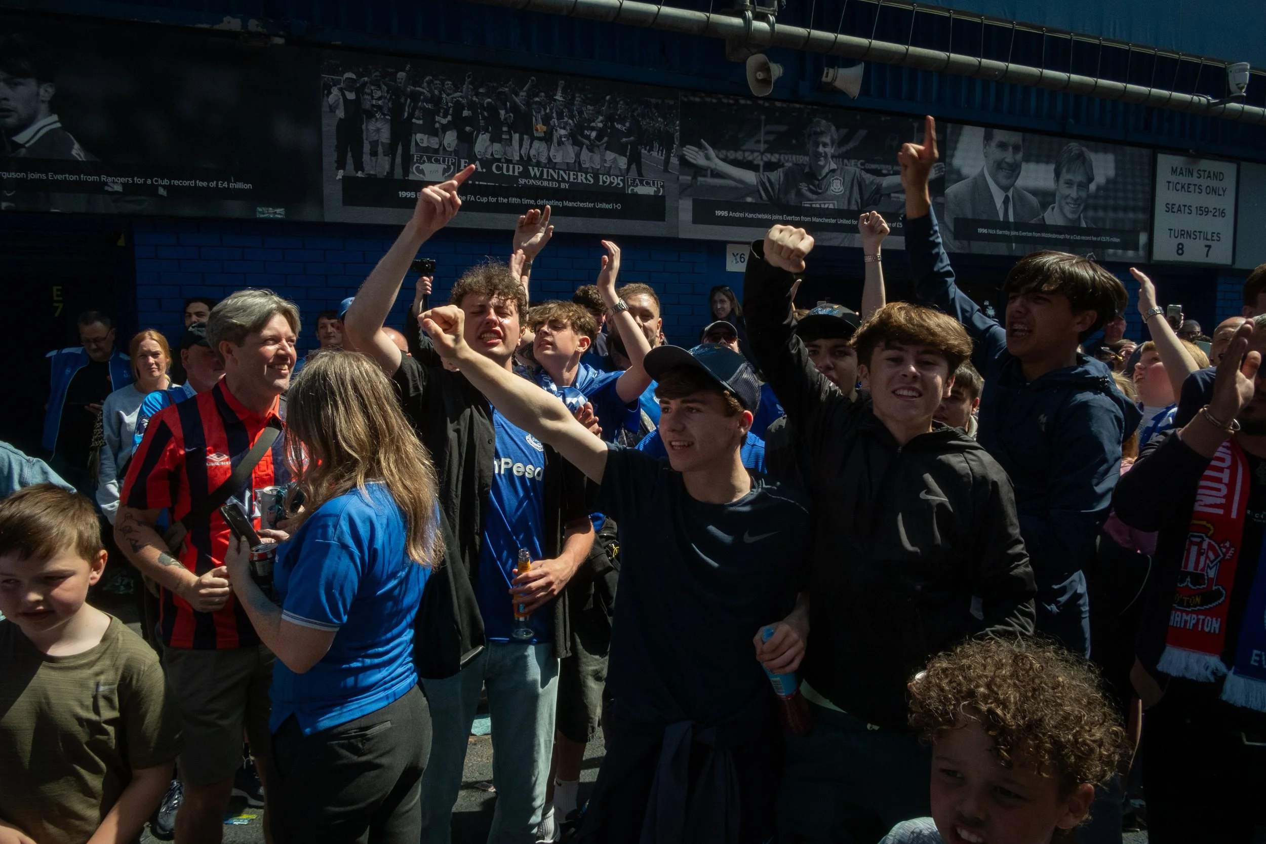 Group of young people celebrating outdoors, some wearing football jerseys, near a stadium entrance with banners and signs.
