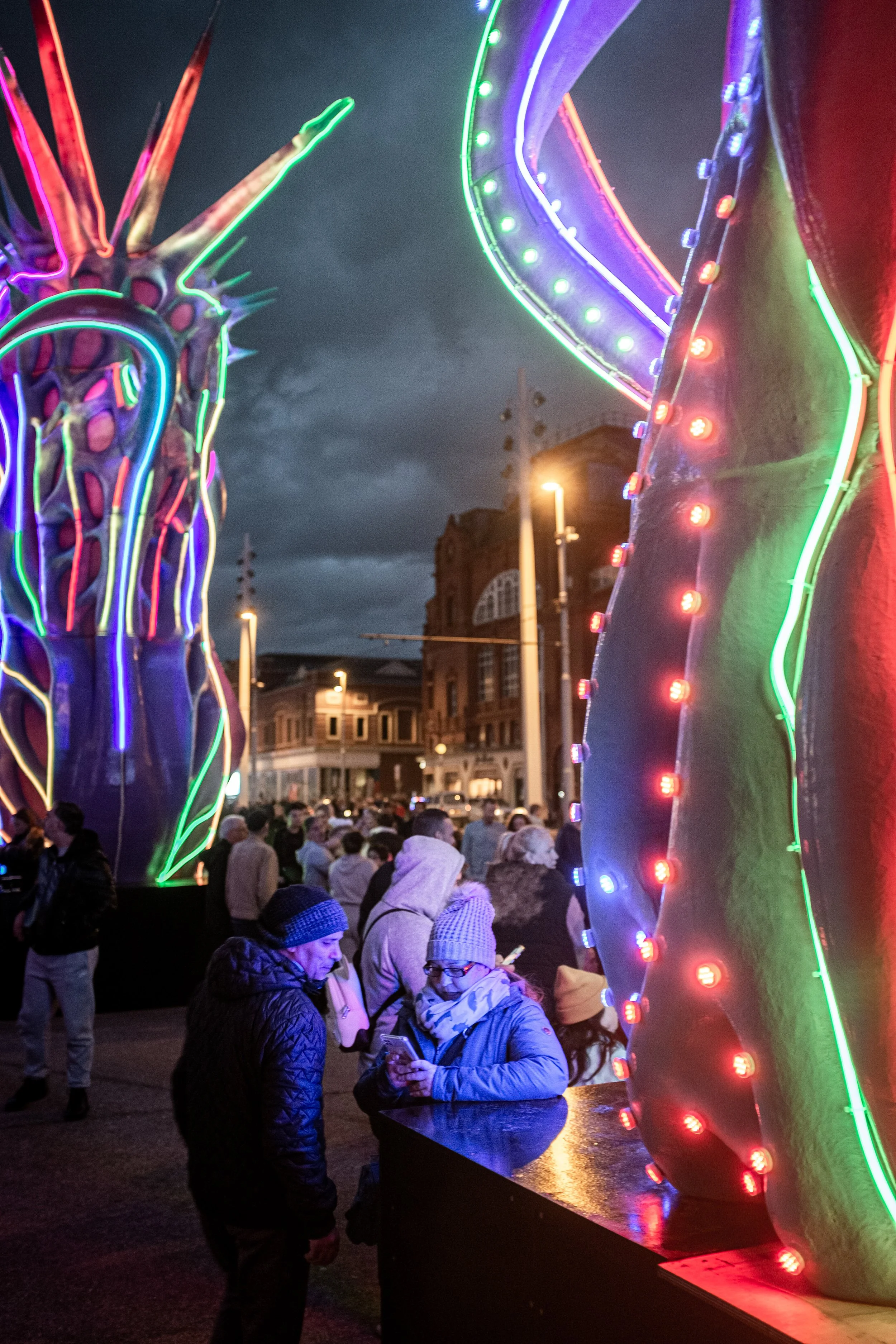 Nighttime scene at a carnival with illuminated large, colorful carnival rides and a crowd of people in jackets and hats