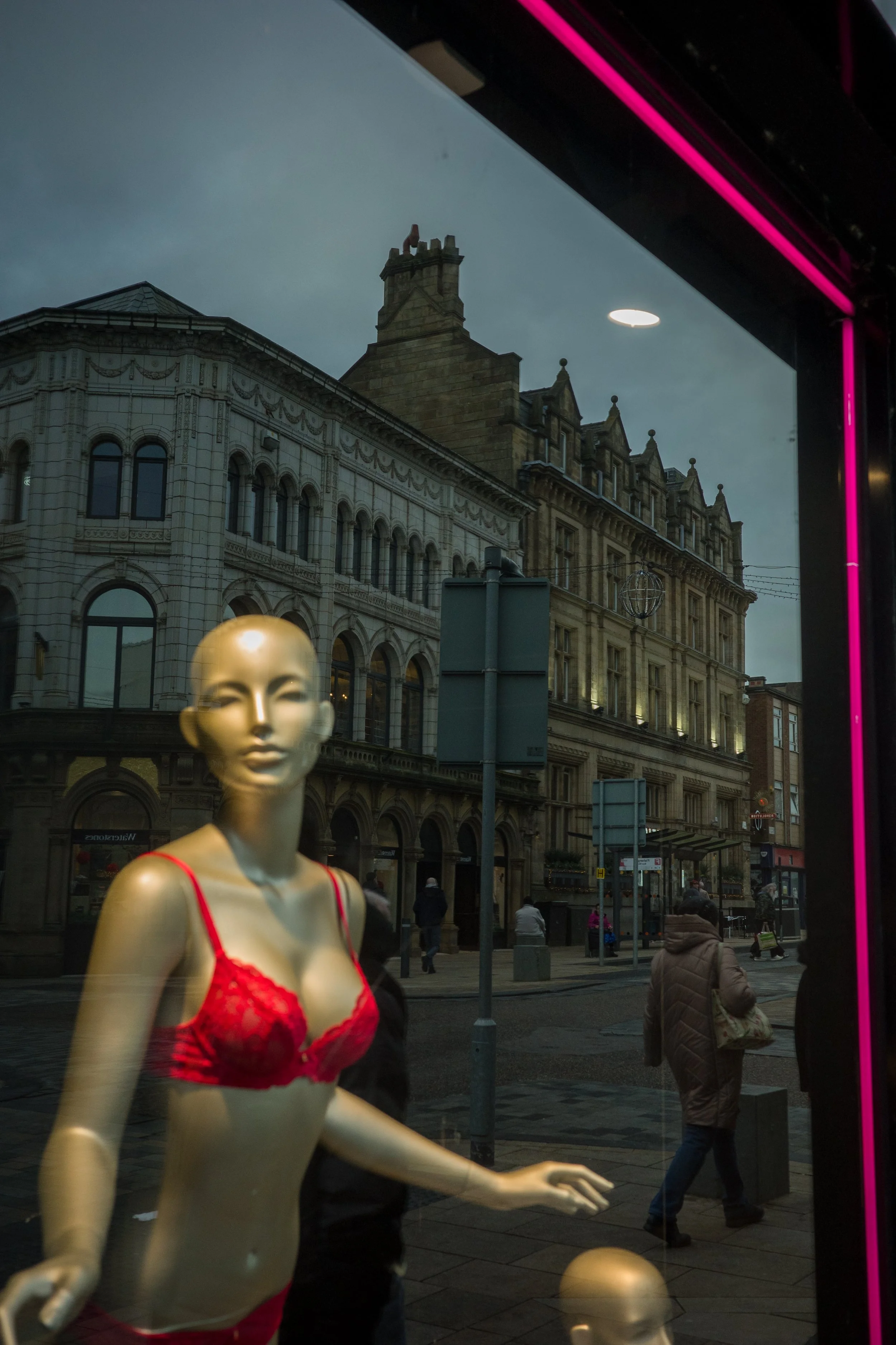 Display window with a mannequin in red lingerie, city street scene reflected in the window with pedestrians and historic buildings.