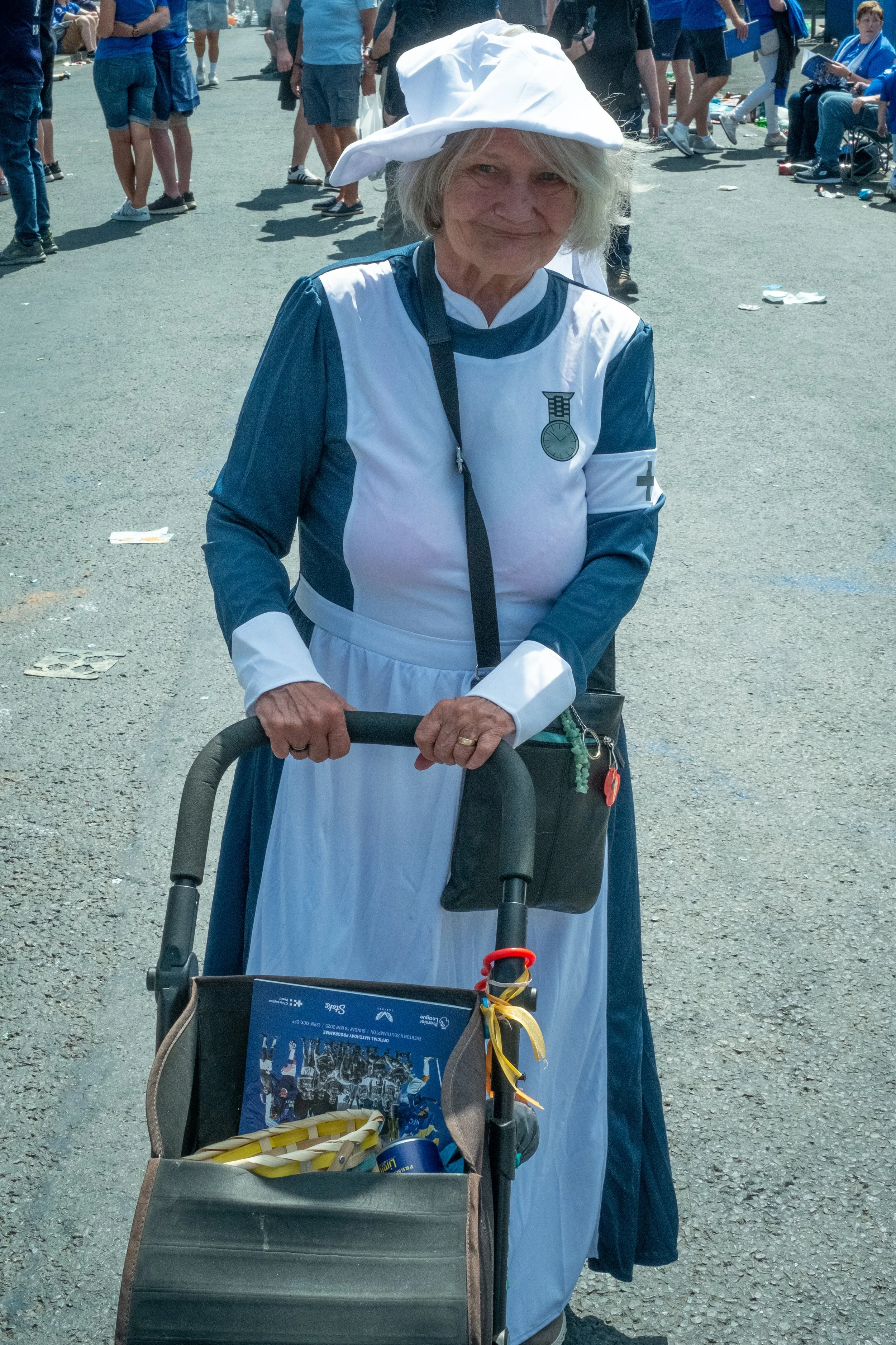 An elderly woman dressed as a nurse, pushing a walker with a decorated basket, at an outdoor event with many people in the background.