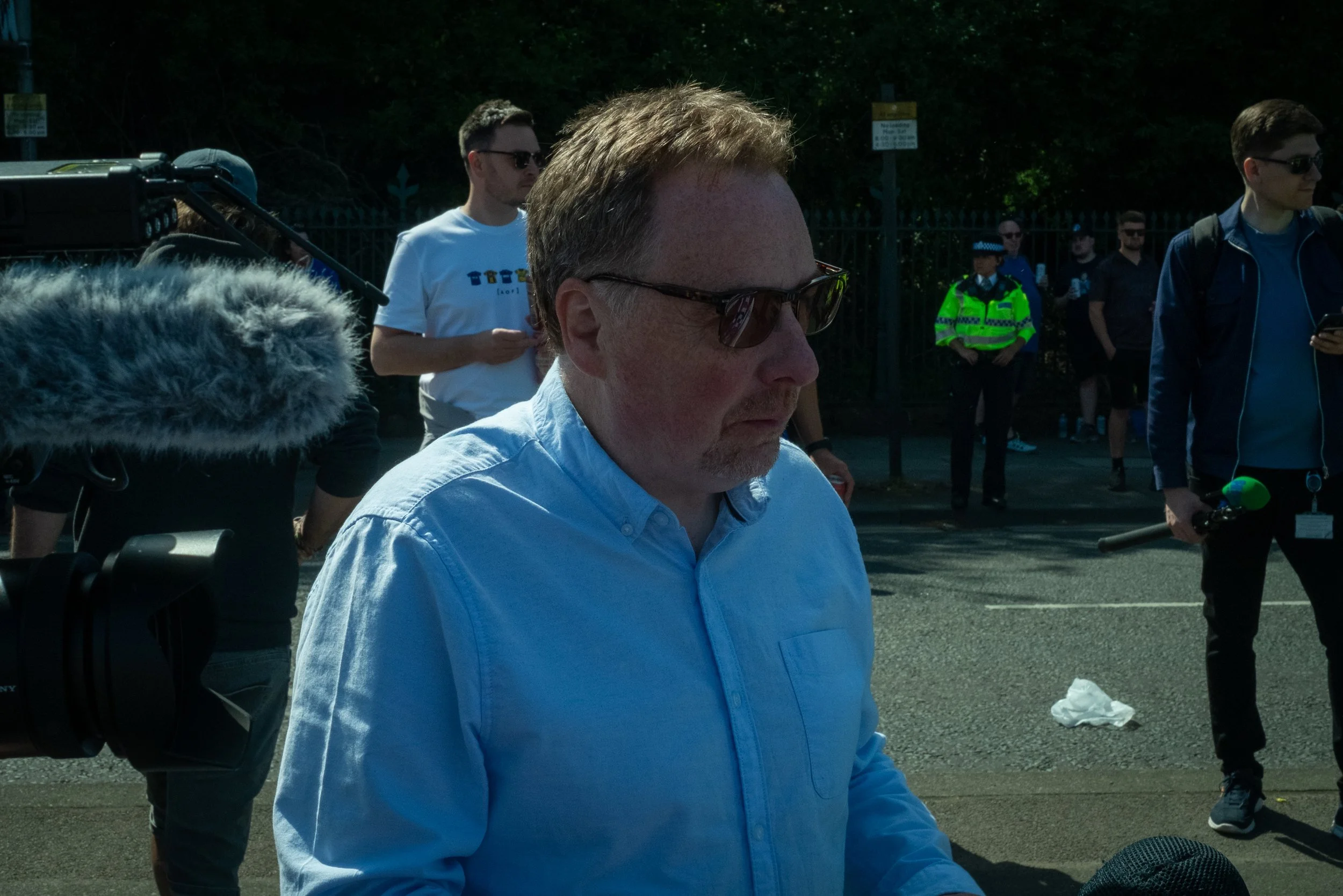 A man with sunglasses and a light blue shirt, surrounded by several people, some holding microphones and cameras, outdoors on a sunny day.