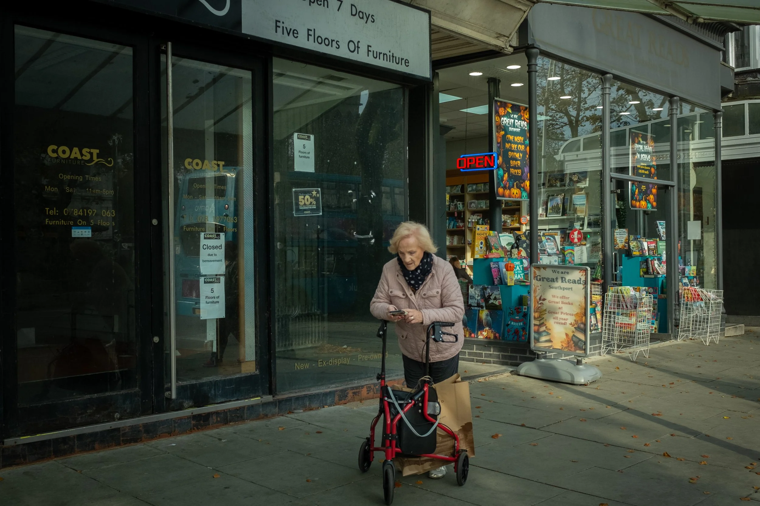 A storefront with a sign that reads 'Five Floors of Furniture' and a neon 'OPEN' sign. An elderly woman with blond hair, wearing a beige jacket and a black scarf with white dots, stands outside with a mobility scooter and a paper bag. There are color