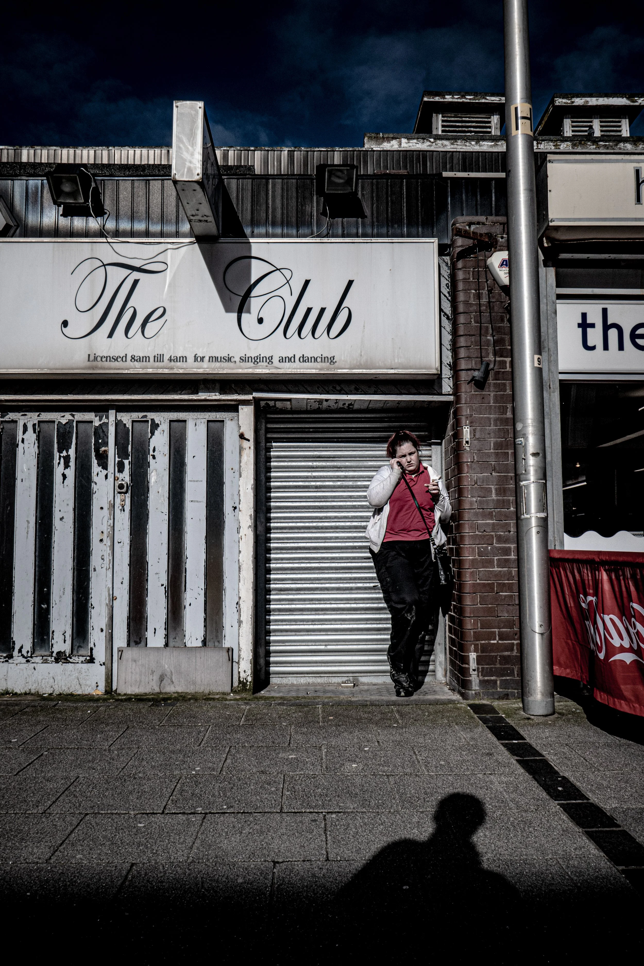 A woman in a red top and white jacket talking on a cellphone on a city sidewalk against a building with a sign that reads 'The Club' and business hours. The sky is dark and cloudy, and there is a shadow of the person taking the photo.
