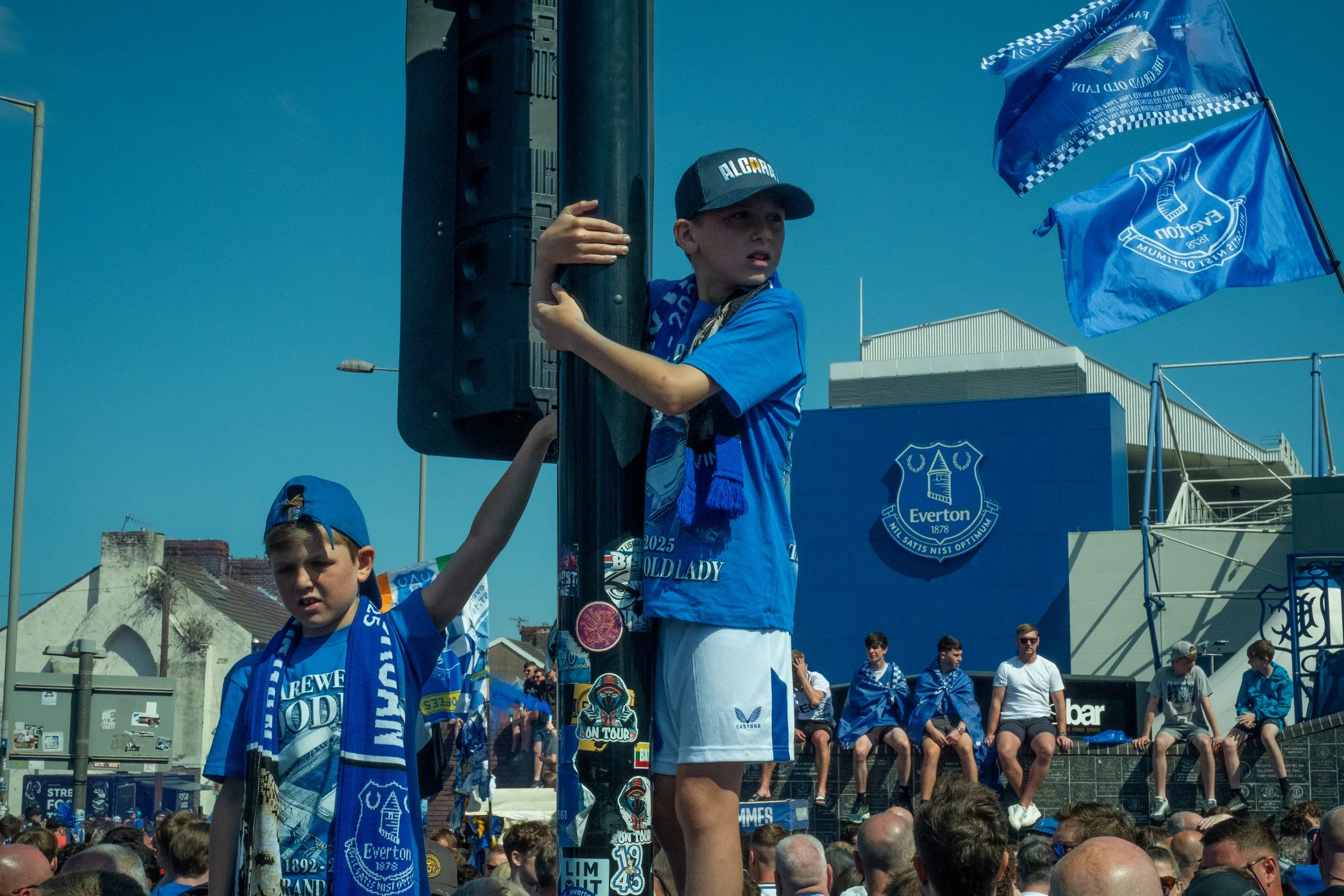 A group of children and adults at a football fan event, with children wearing Everton football team jerseys, scarves, and hats, gathered outdoors on a sunny day.