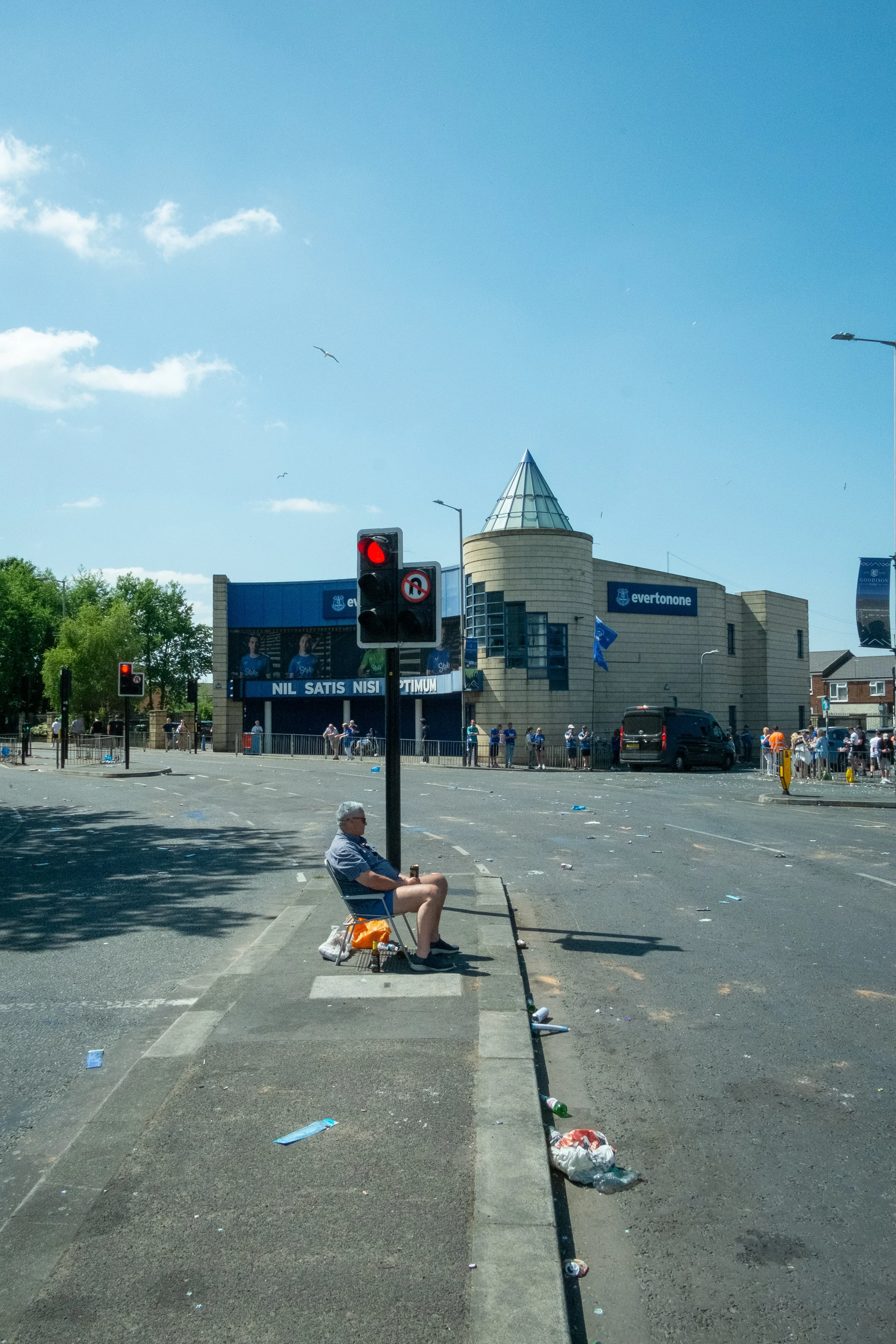 A man sitting on a folding chair on a sidewalk curb in front of a traffic signal, with litter scattered around. In the background, there is a stadium with a sign reading 'evertonone' and a crowd outside.