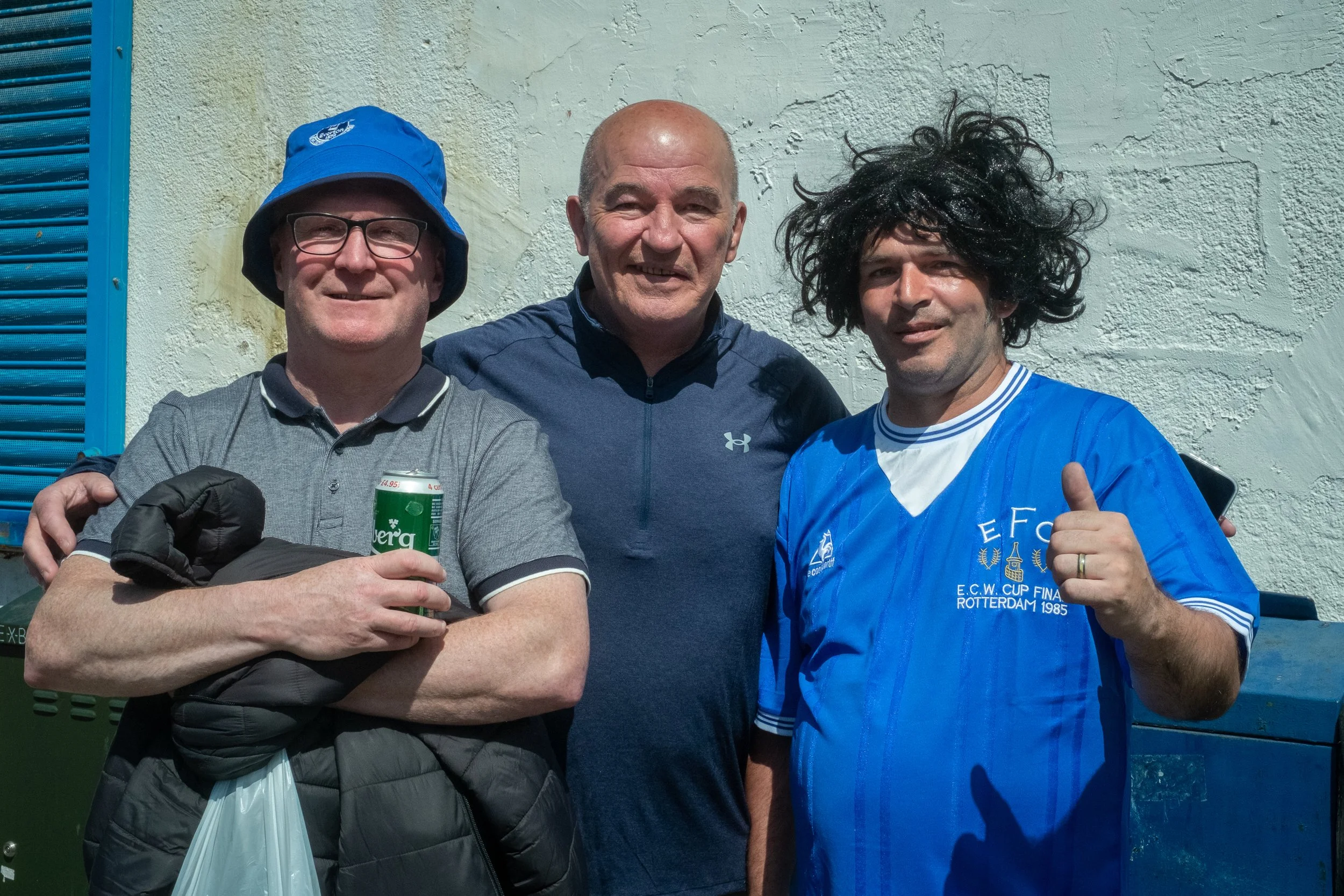 Three men standing together outside against a white wall, smiling at the camera. The man on the left is wearing glasses, a blue bucket hat, a gray polo shirt, and holding a can of beer. The man in the middle is bald, wearing a dark blue Under Armour 