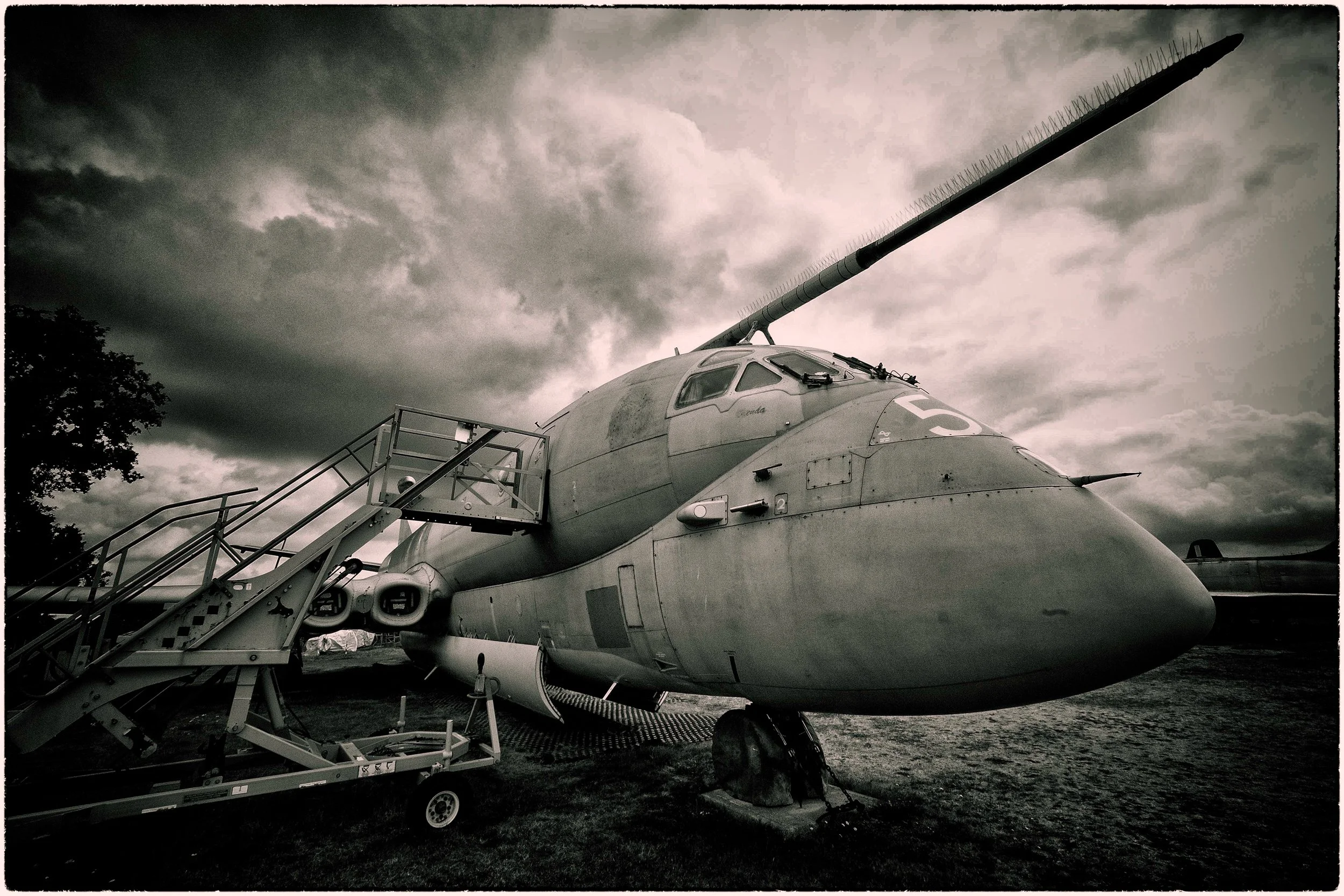 A military fighter jet with a long missile attached to its nose, parked outdoors under a cloudy sky, with a staircase leading up to the cockpit.