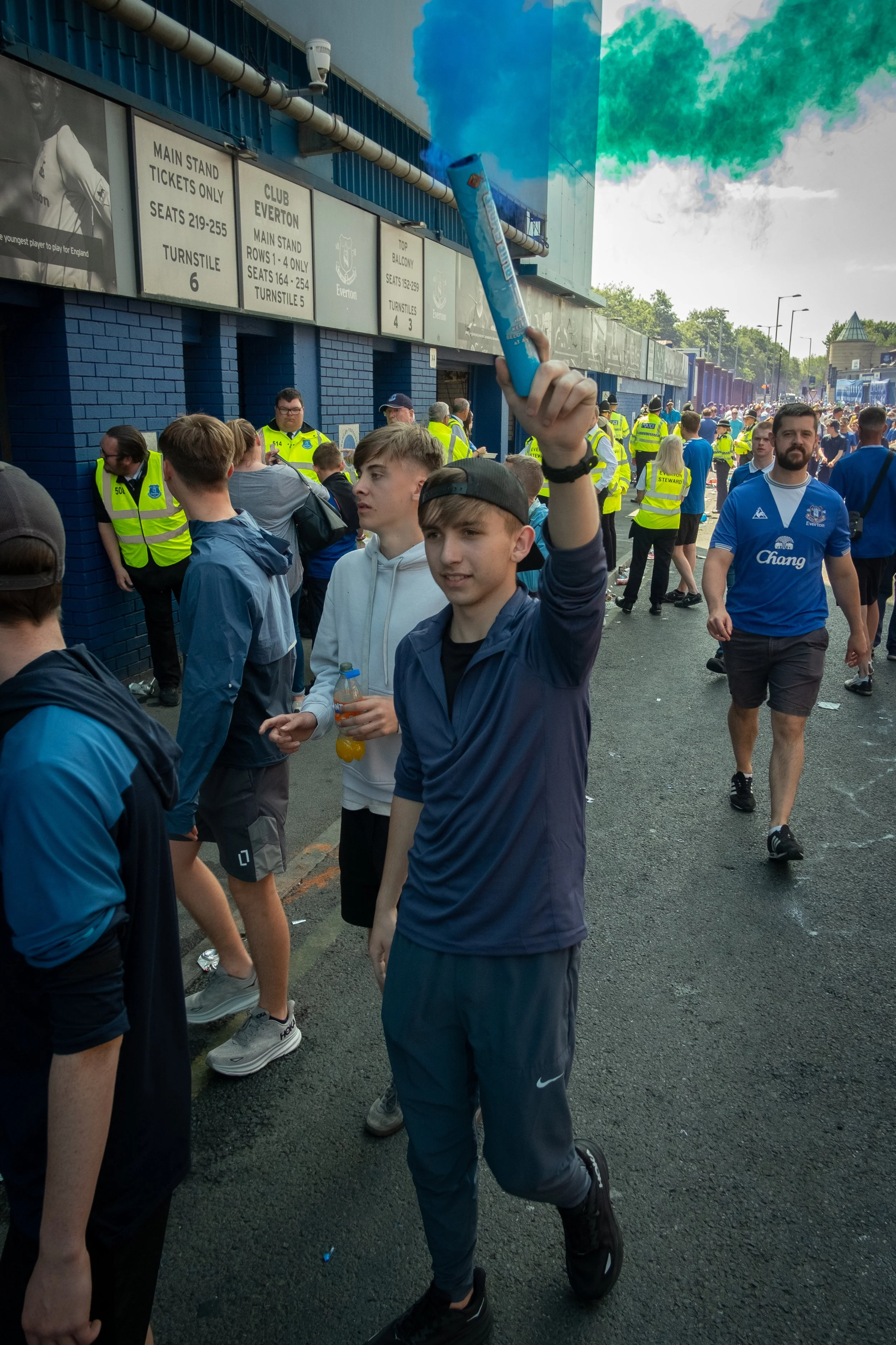 Crowd of fans outside a football stadium, with one young man in dark athletic clothing holding a blue smoke flare.