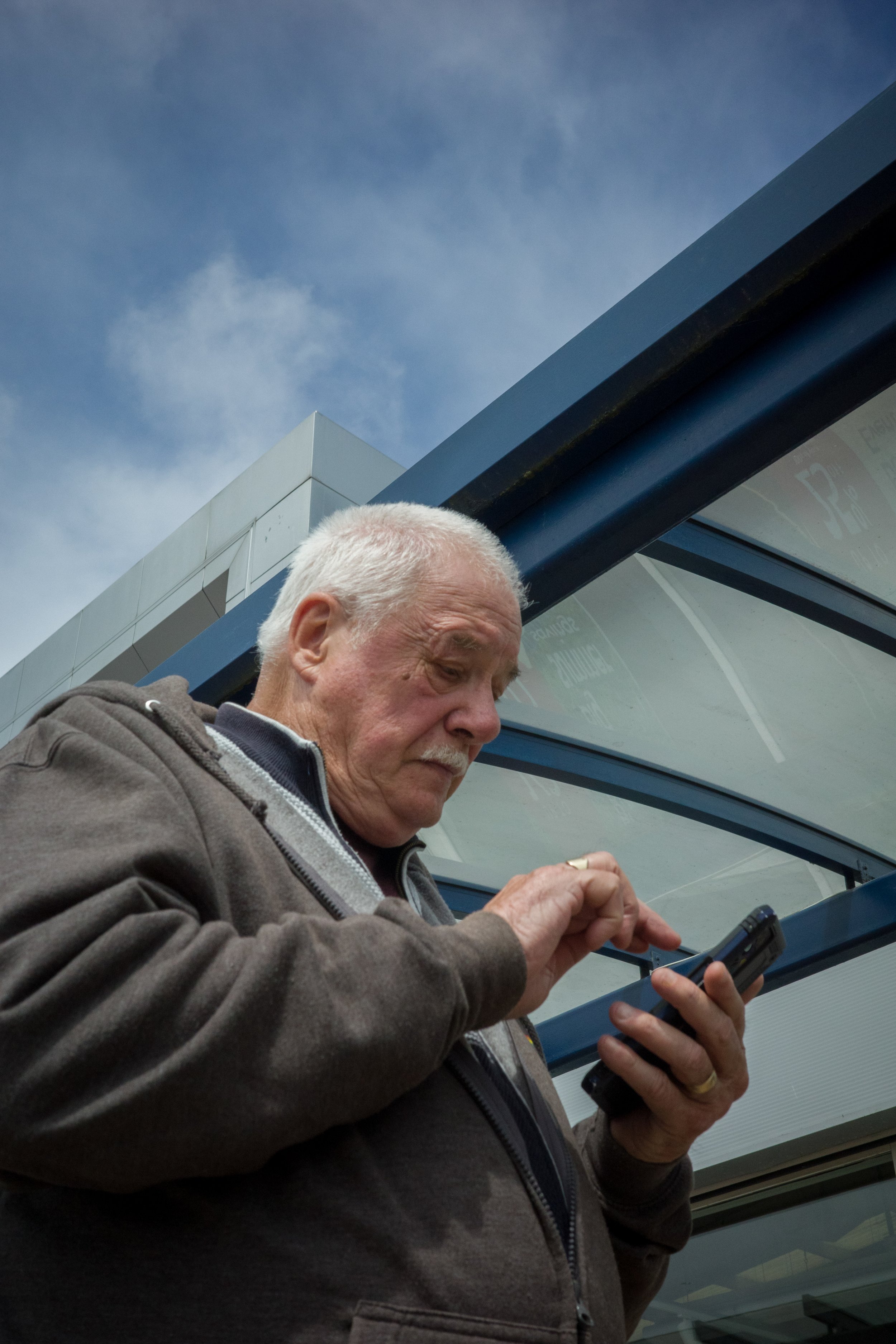 An elderly man with white hair and a gray hoodie looking at a smartphone at a bus stop with a modern building and blue sky in the background.