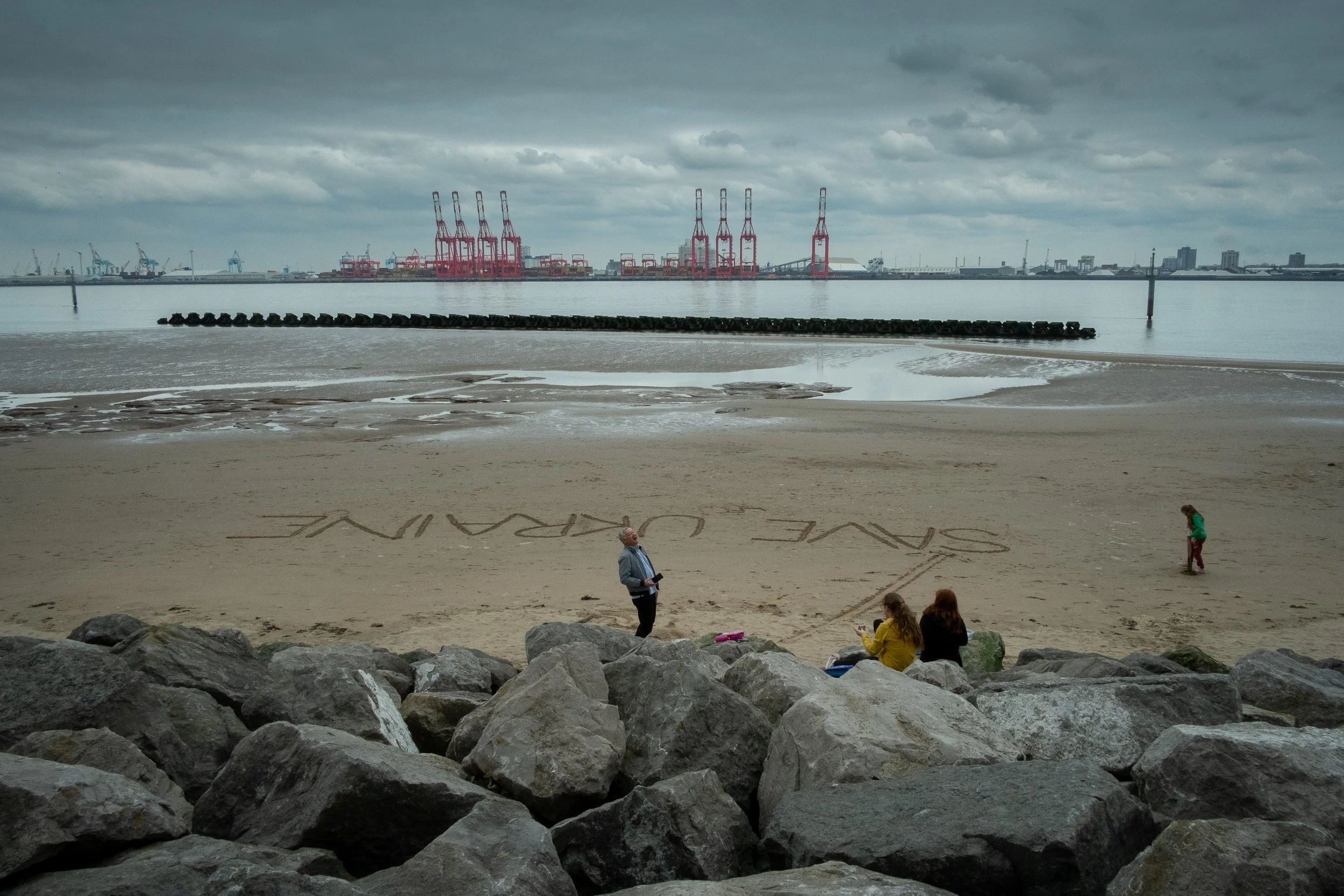 A beach scene with a cloudy sky, displaying a large body of water, rocks in the foreground, and industrial cranes in the distance. There are three people on the beach, two sitting on rocks, one standing with a camera, and a child walking on the sand.
