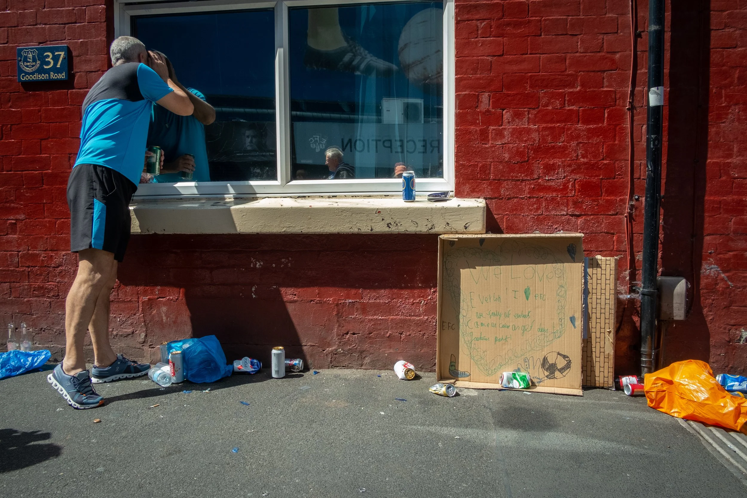 A man in athletic clothes embraces someone through a window ledge, with litter and a cardboard sign on the sidewalk outside a red brick building.