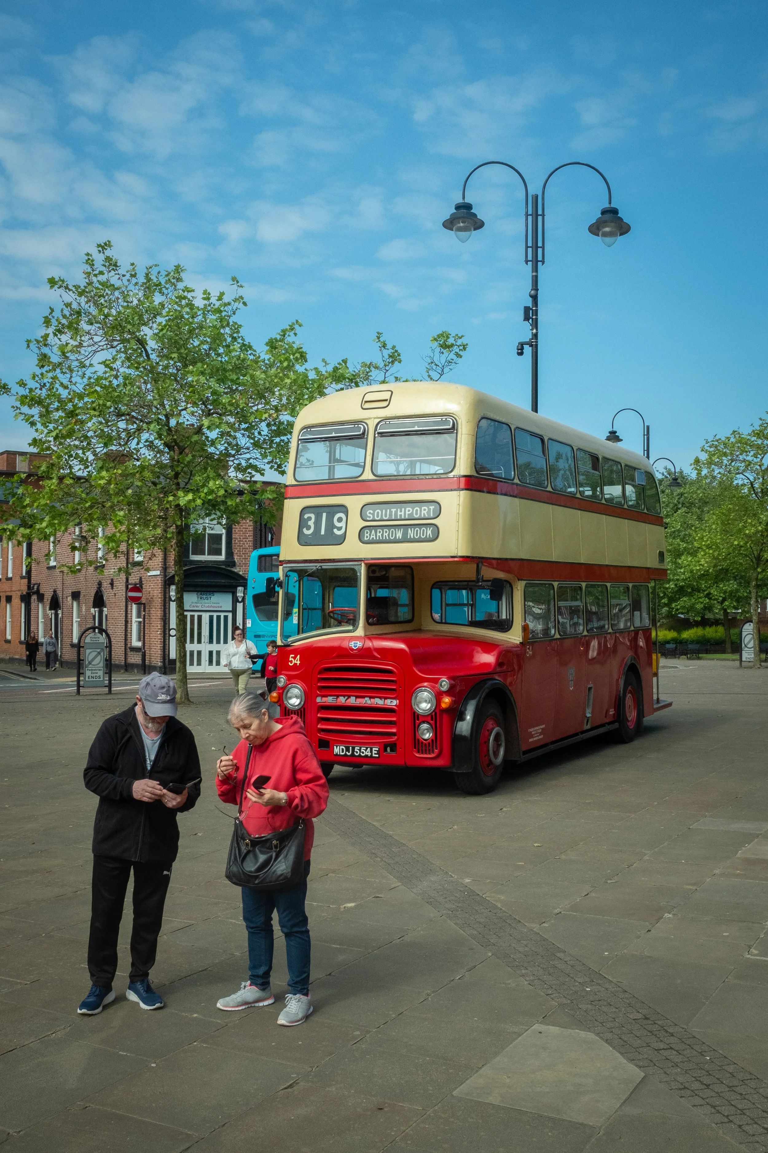 An old double-decker bus painted in red and cream, with the destinations Southport and Barrow Nook displayed on the front, parked on a pavement. In the foreground, two elderly people are looking at their phones, and a group of people are visible walk