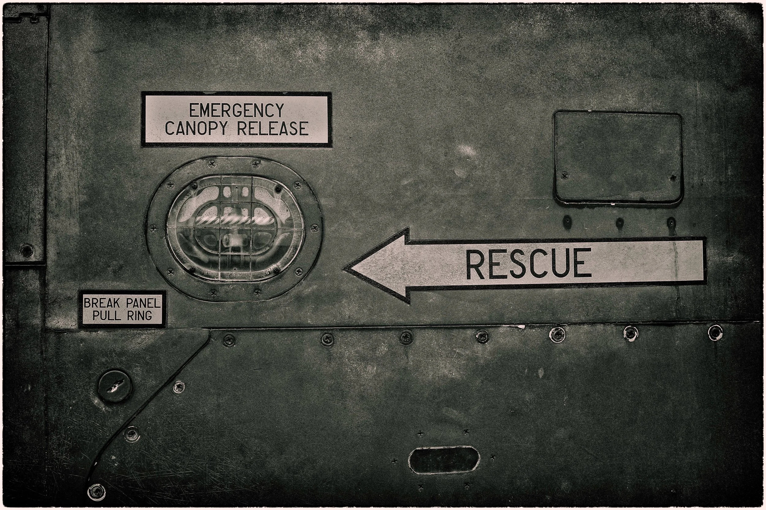 A metal panel with an emergency canopy release label, a rescue arrow pointing to the right, a circular vent with a grid cover, and a break panel pull ring labeled in small print.