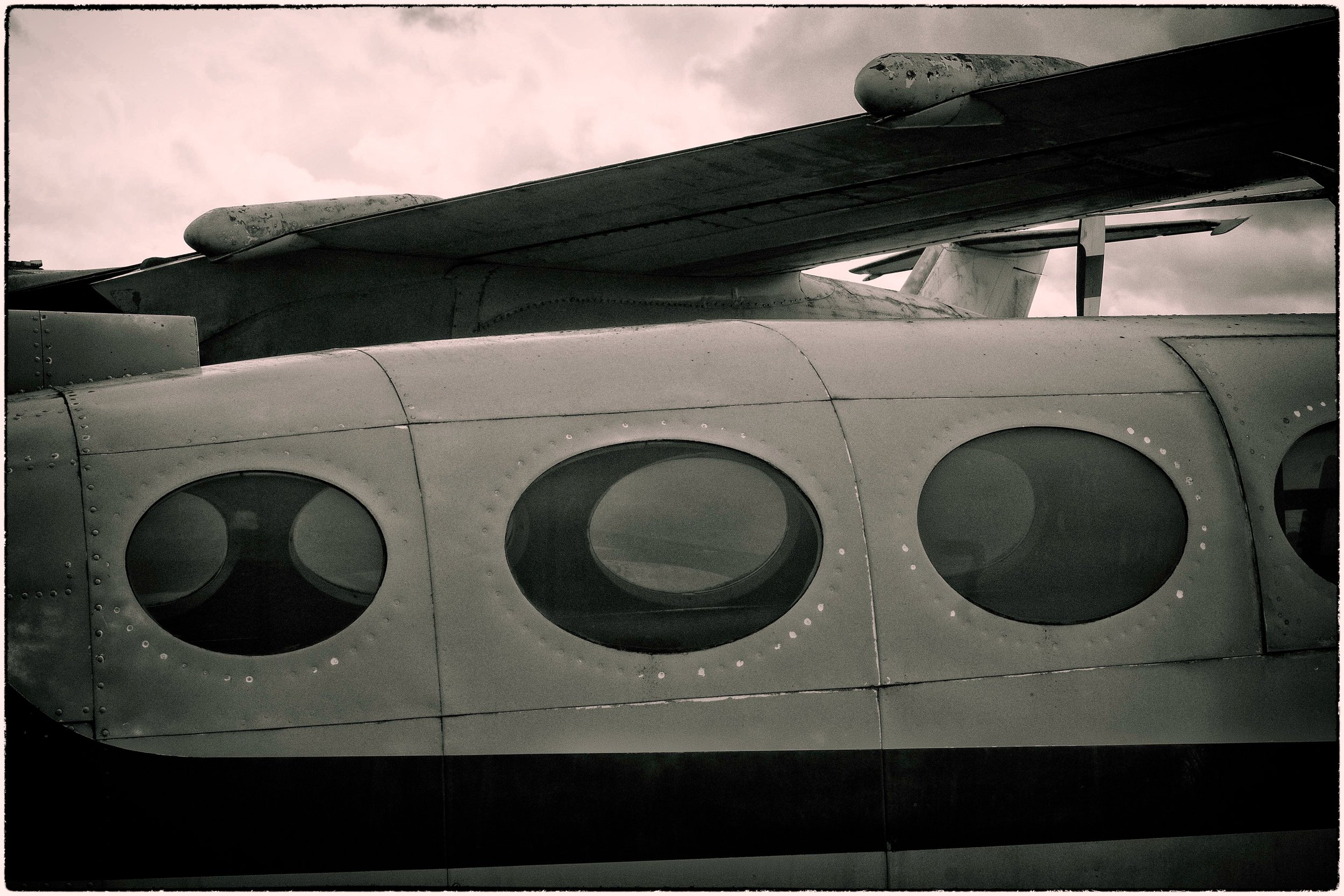 Close-up of an aircraft wing with a turbo fan engine and fuselage with round windows, under a cloudy sky.