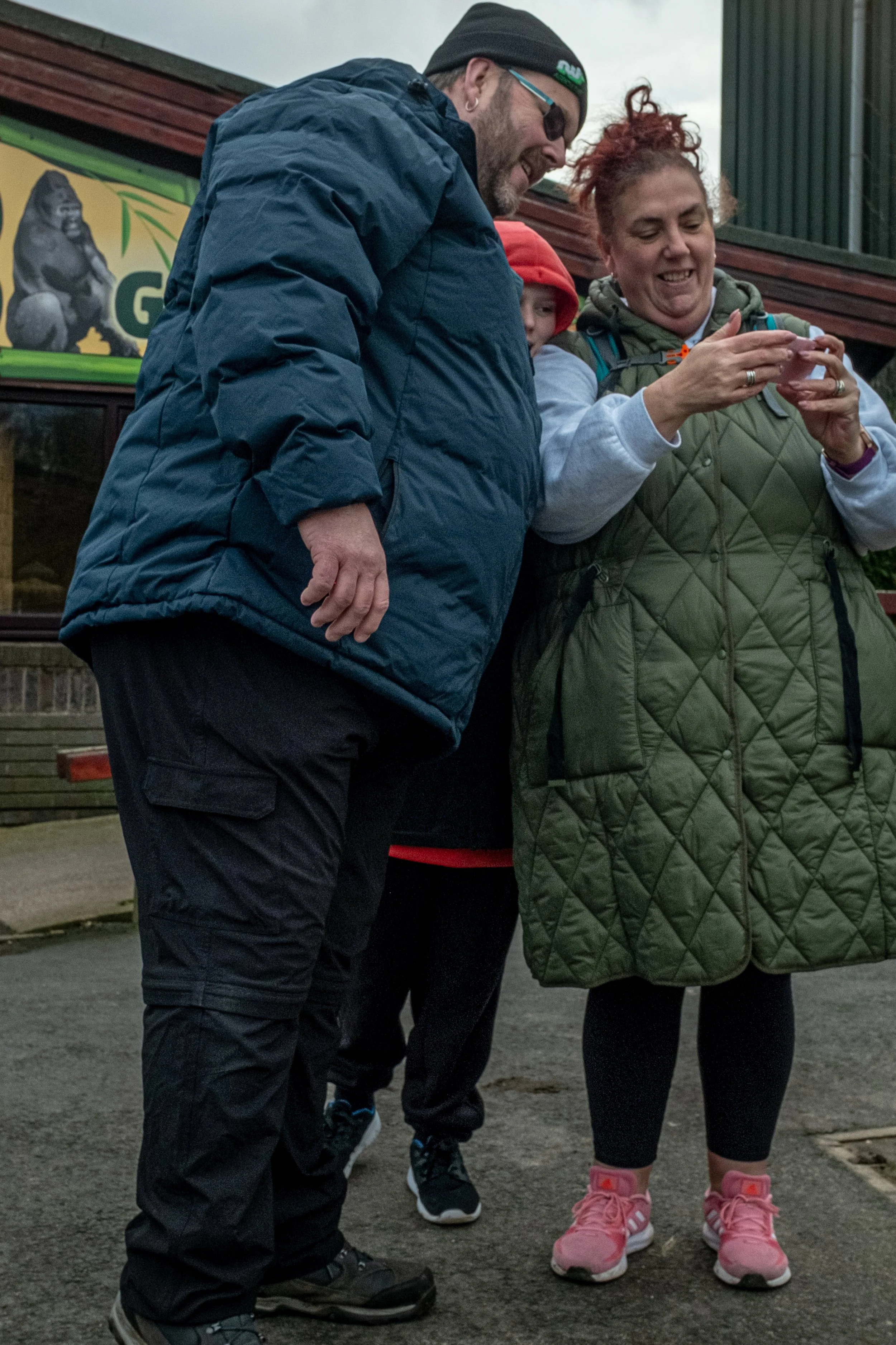 Three people are standing close together outdoors, looking down at something. They appear to be in casual clothing with jackets. One woman is showing an object to a man and a child. The background features a building with a sign showing a gorilla.