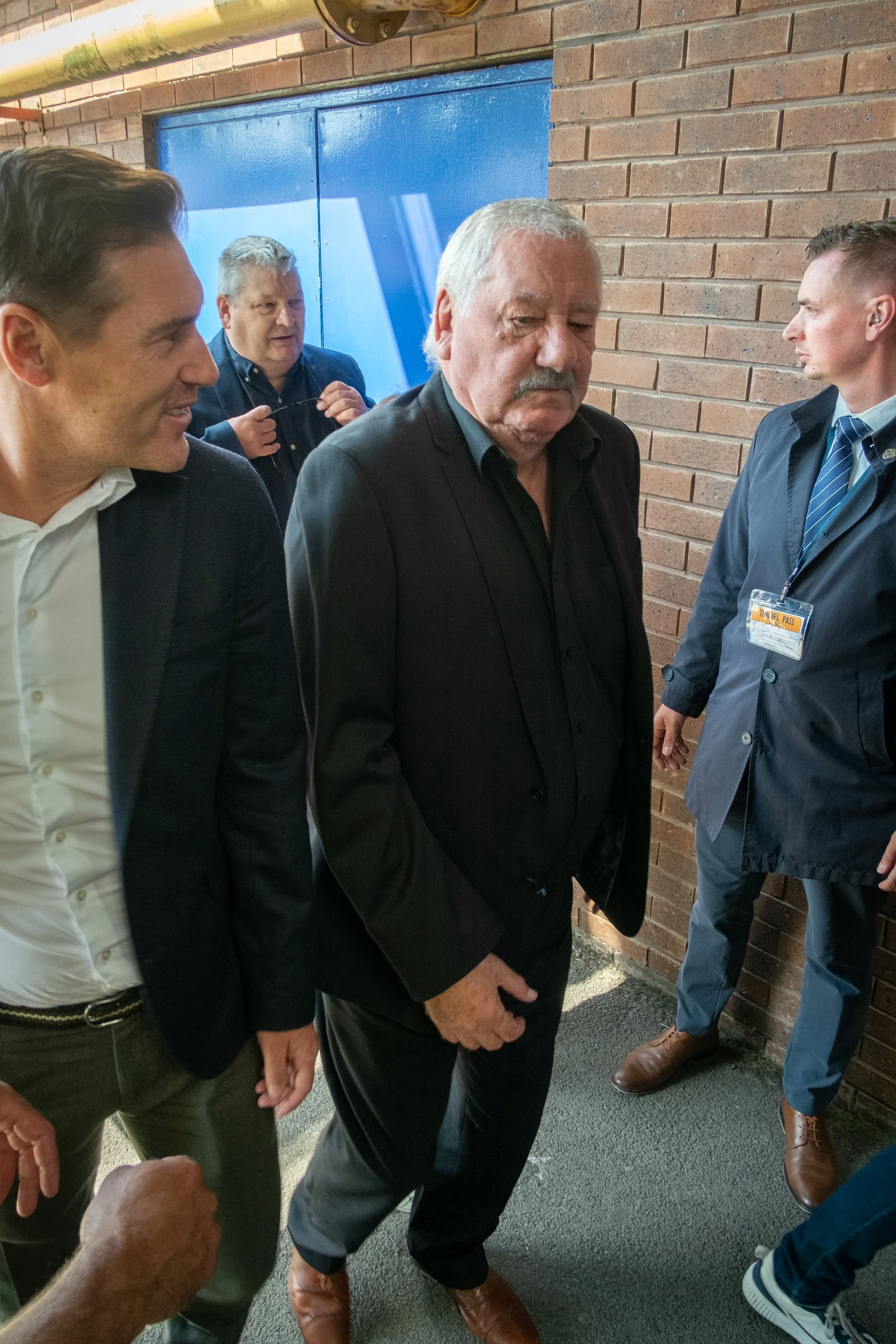 A group of men standing and talking in a hallway with a brick wall and a blue door in the background. One man in the center appears to be upset or serious, with others around him engaging in conversation.