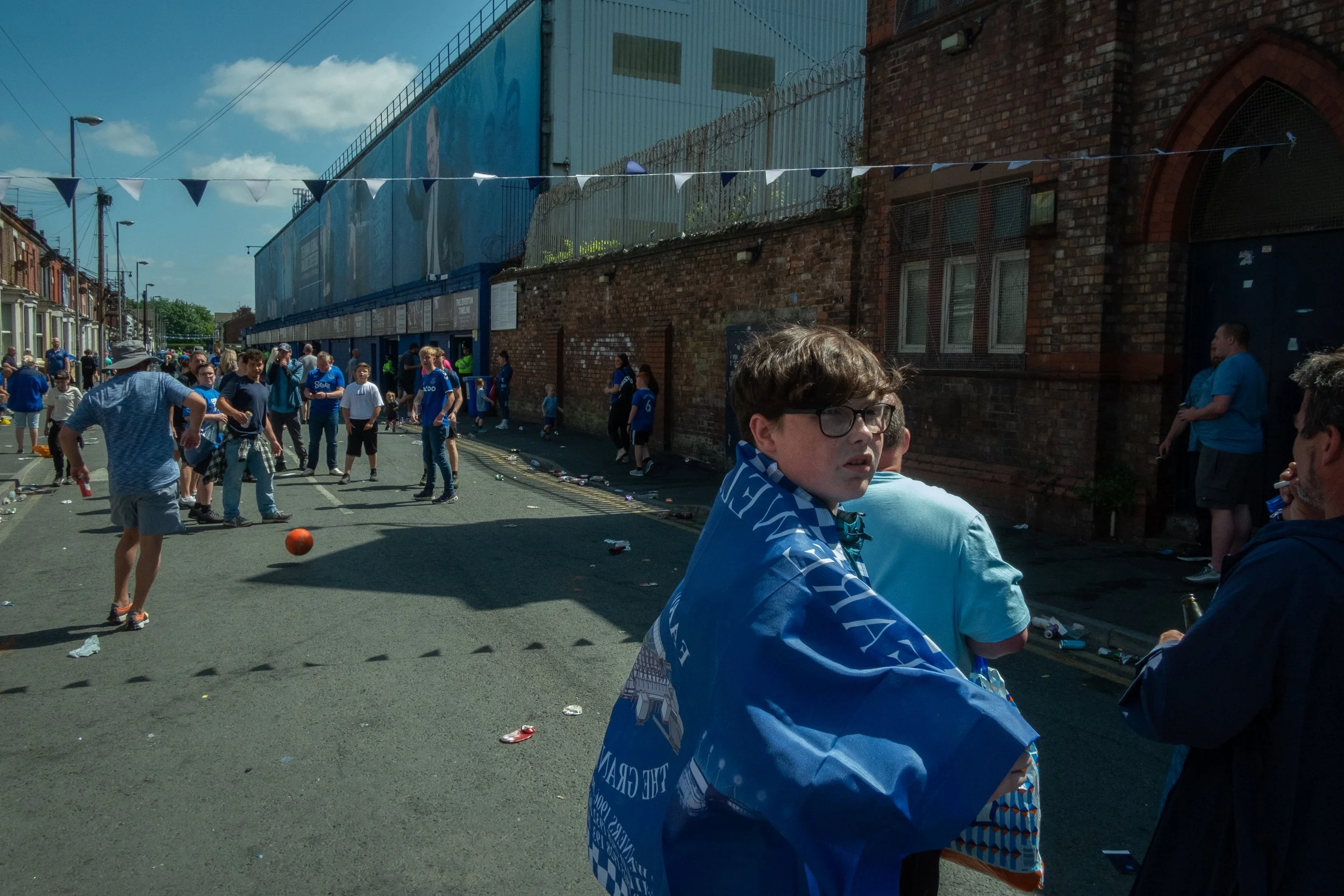People gathering on a street after what appears to be a sports event, with some wearing blue jerseys and flags, and a young man in the foreground wearing glasses and a blue scarf.